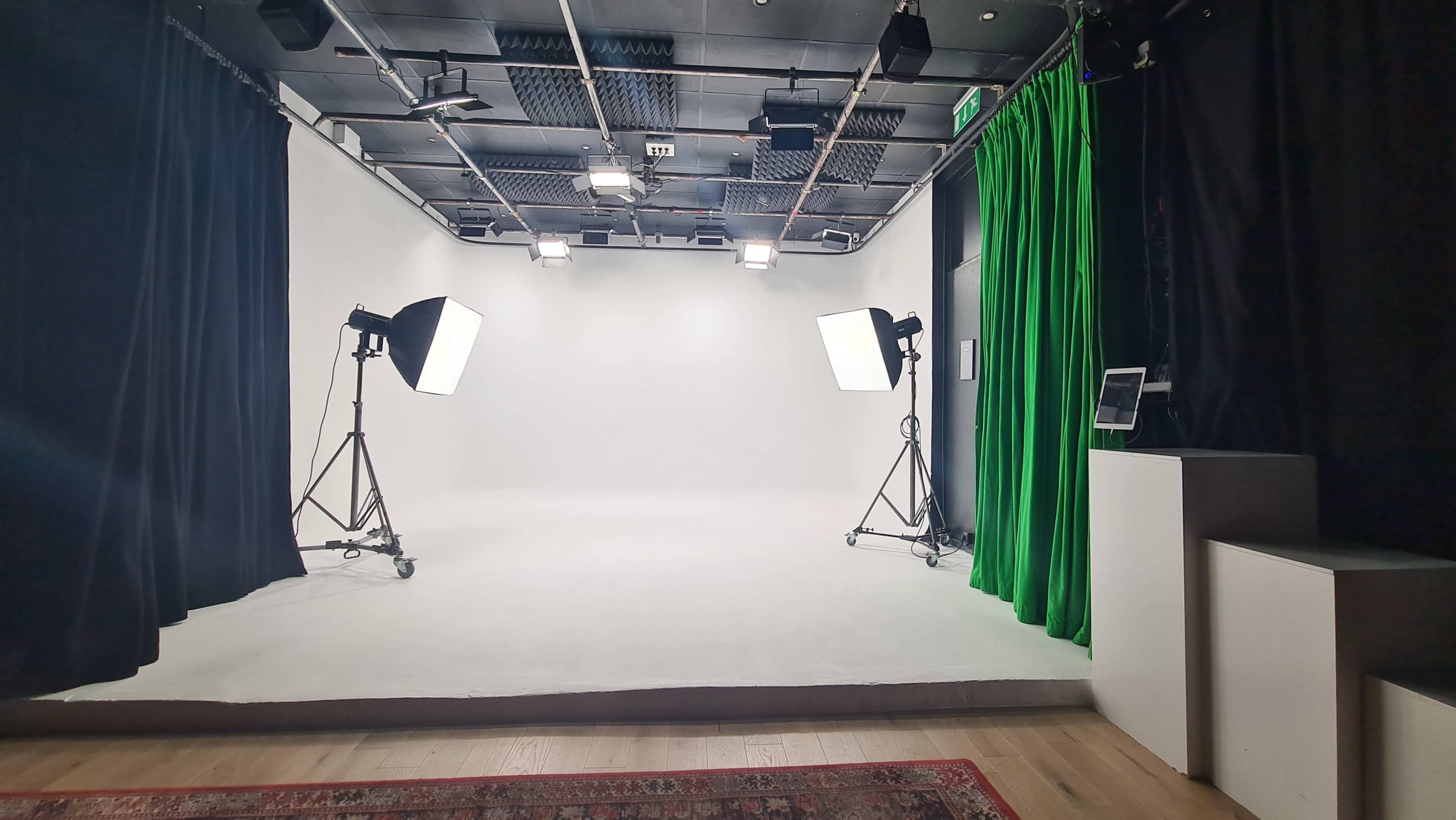 The image shows a photography studio with a plain white backdrop, two light setups on stands, and a green curtain on one side.