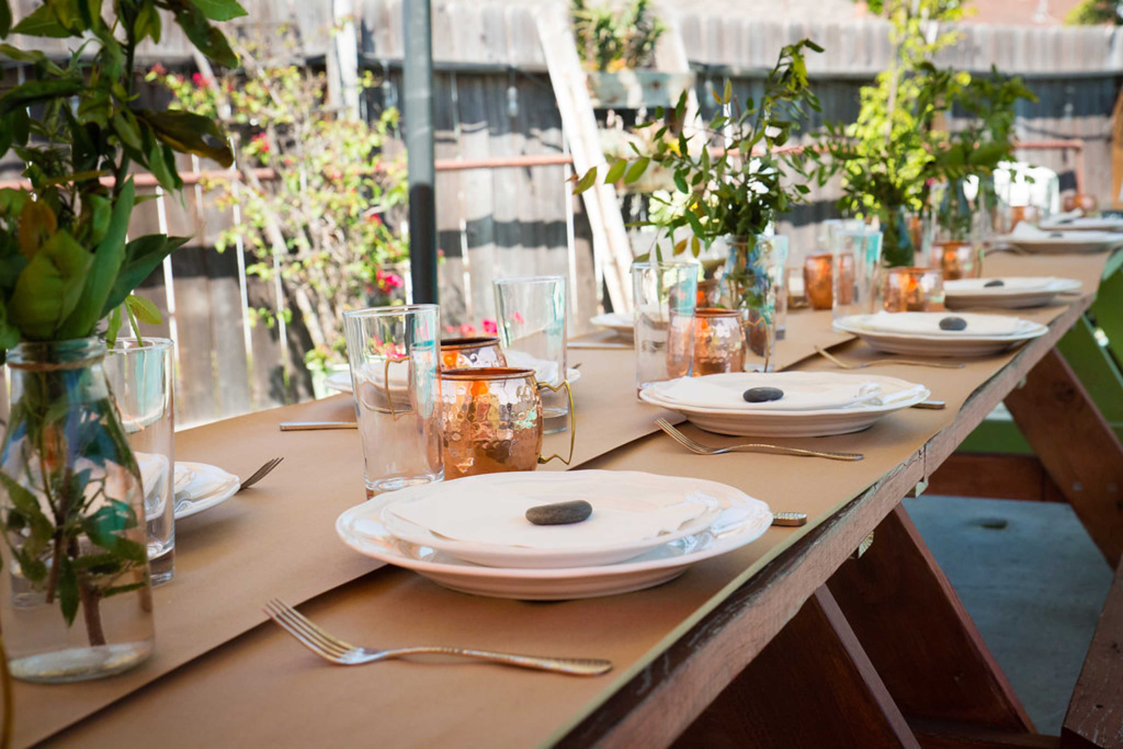 A long wooden table is set with white plates, glasses of water, and vases of green plants, all placed on brown paper.