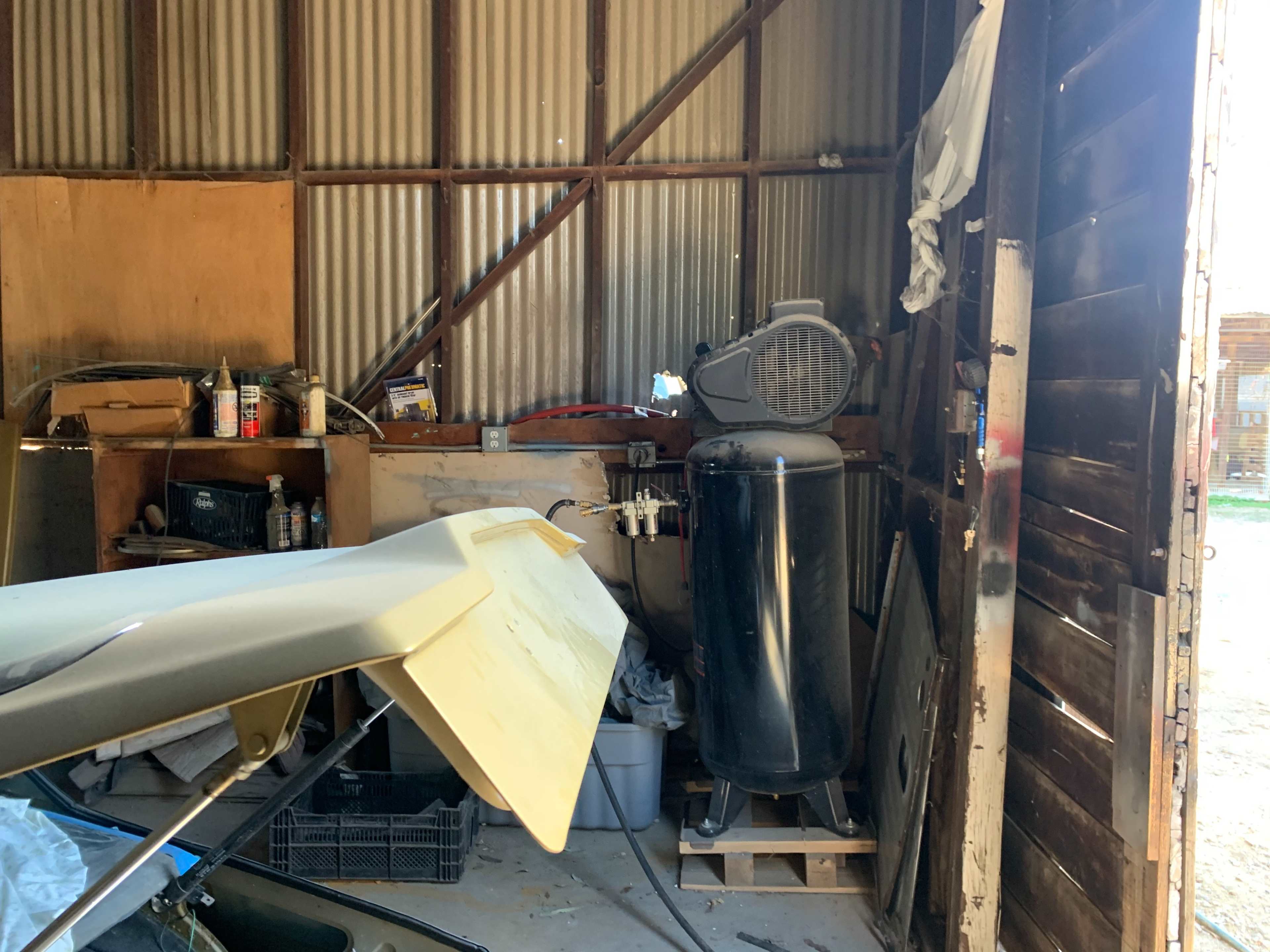 A cluttered shed interior featuring a black air compressor, various tools, and a partial view of a vehicle part.