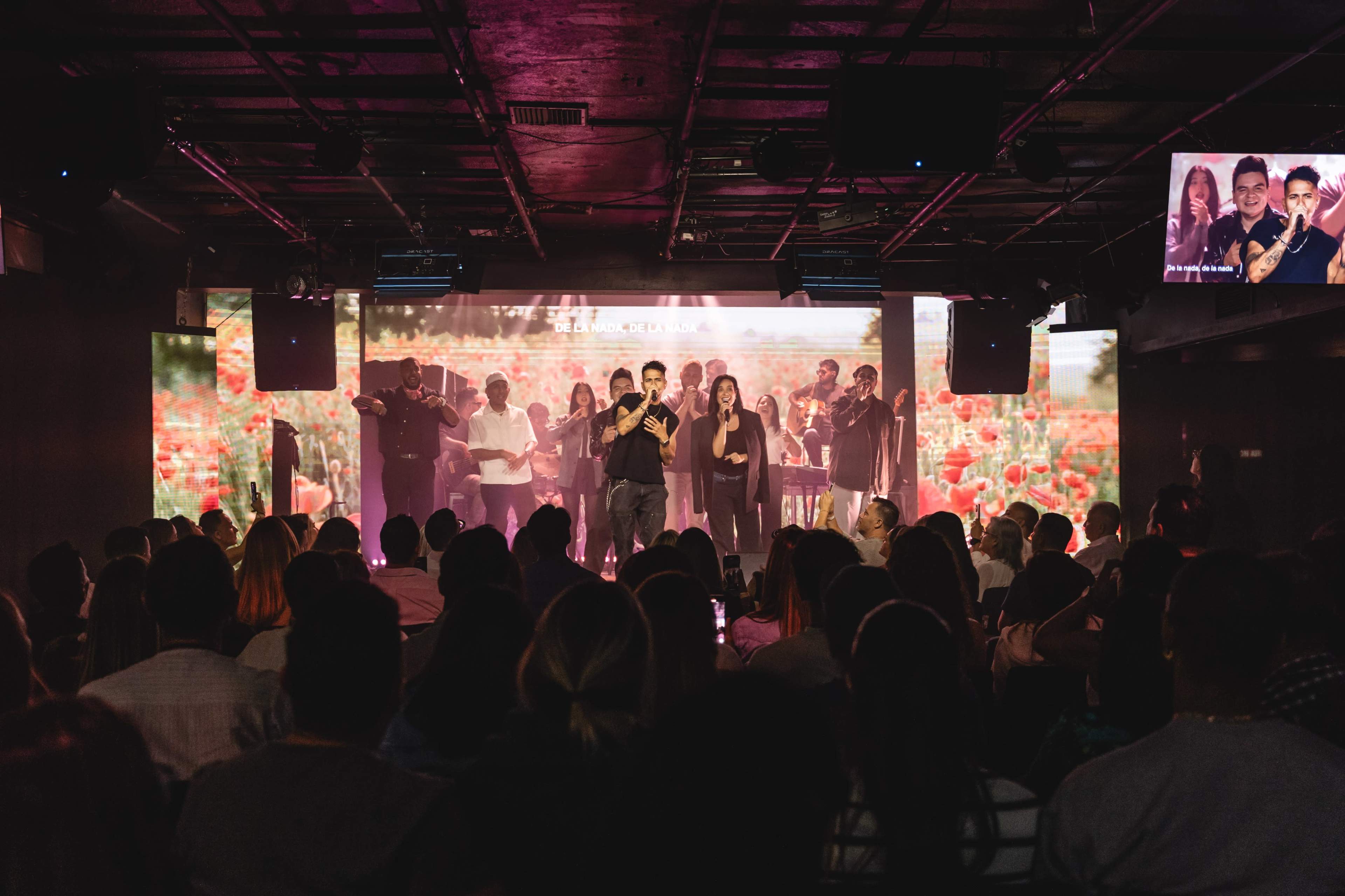 A group of performers stands on stage in front of a large screen displaying a scenic background, while an audience watches from below.