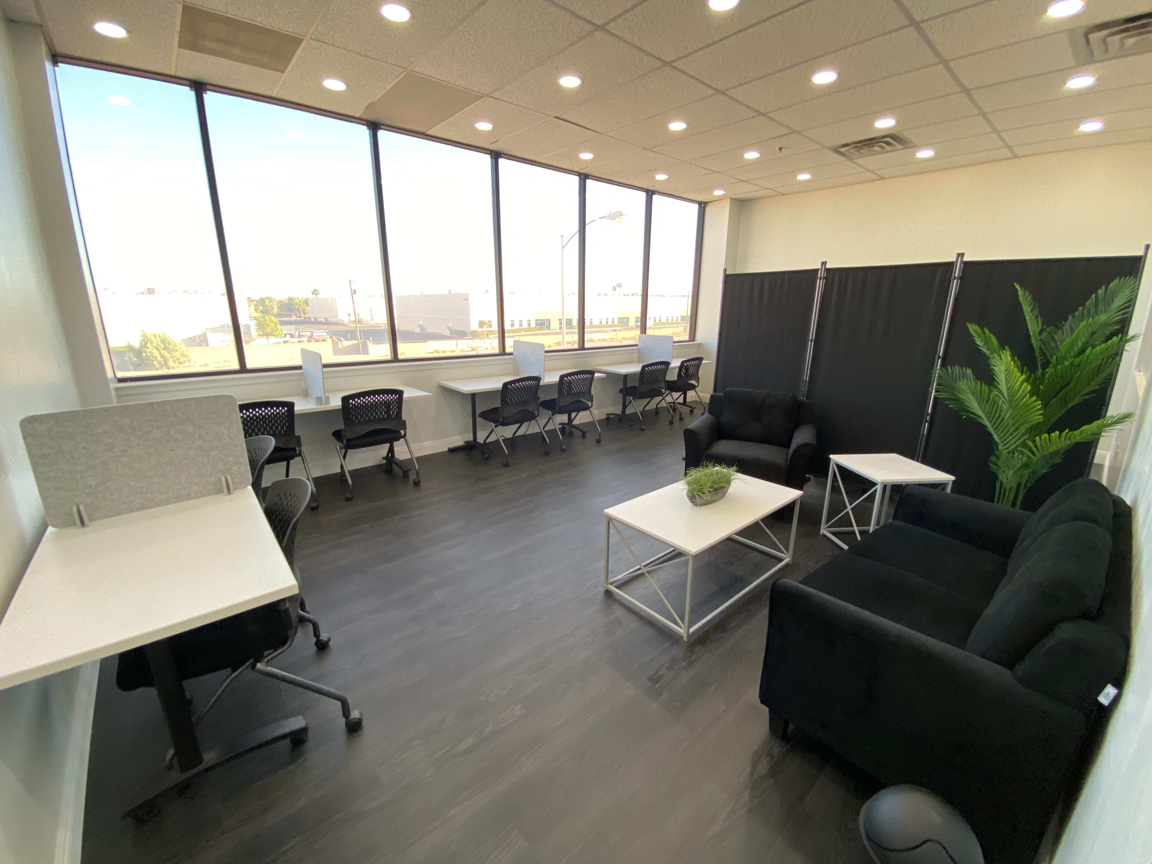 The image shows a bright, modern office space with several desks, chairs, and a seating area featuring two black couches and a table.