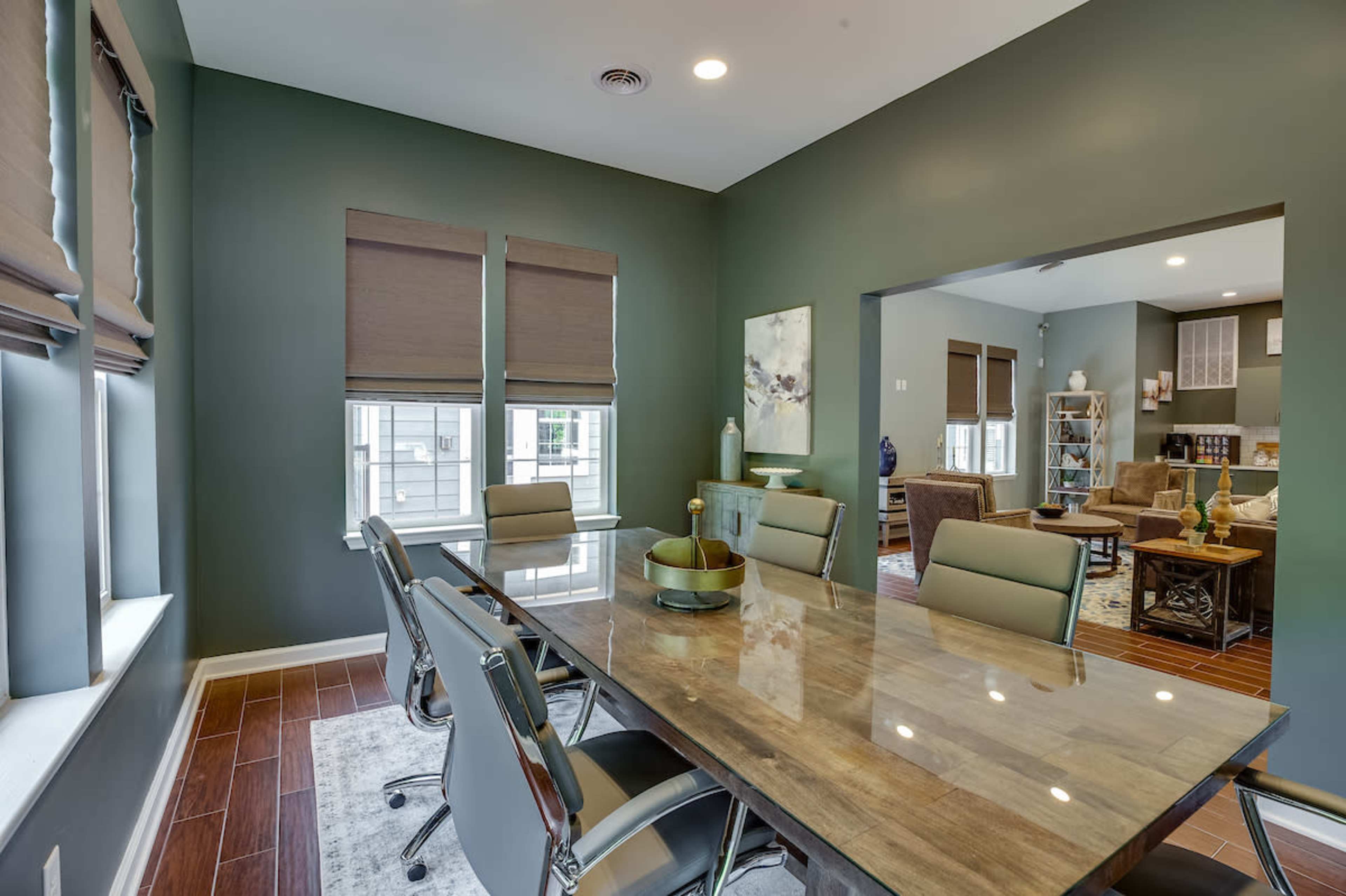 A modern meeting room features a glass table surrounded by gray chairs, with large windows covered by brown blinds and green walls.