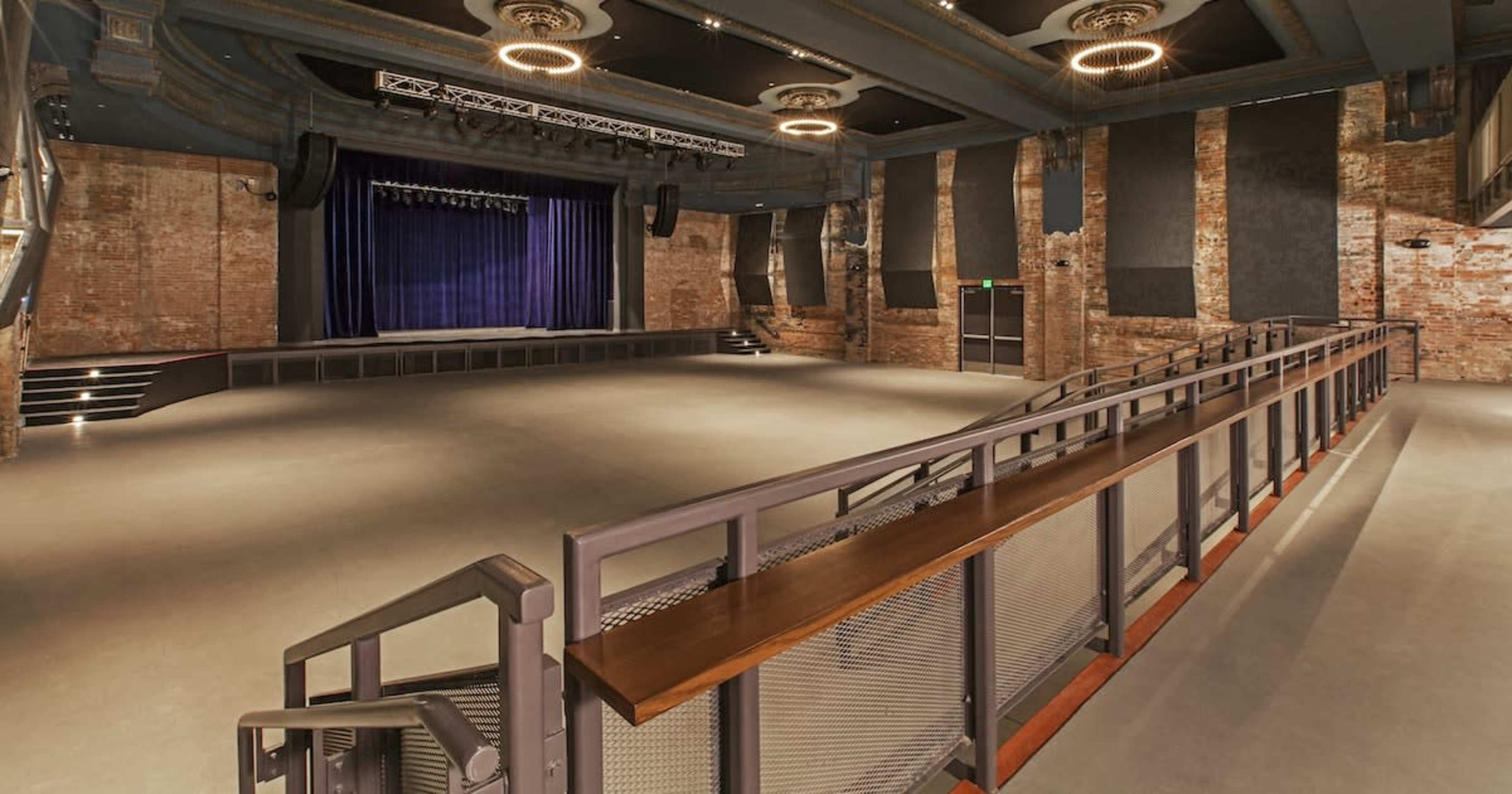 An empty auditorium with a wooden railing overlooking a stage surrounded by exposed brick walls and spotlights.