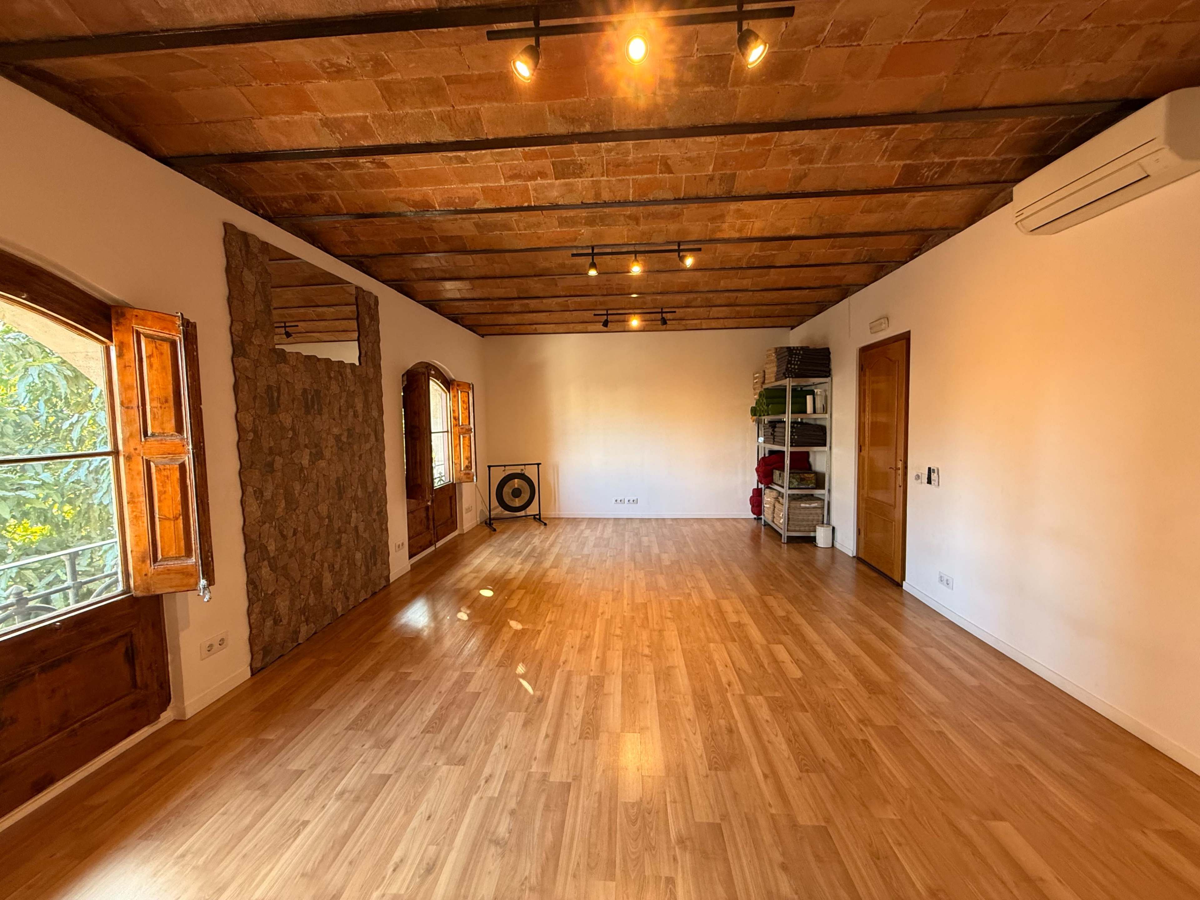 The image shows a spacious room with wooden flooring, exposed ceiling beams, and large windows that allow natural light to enter.