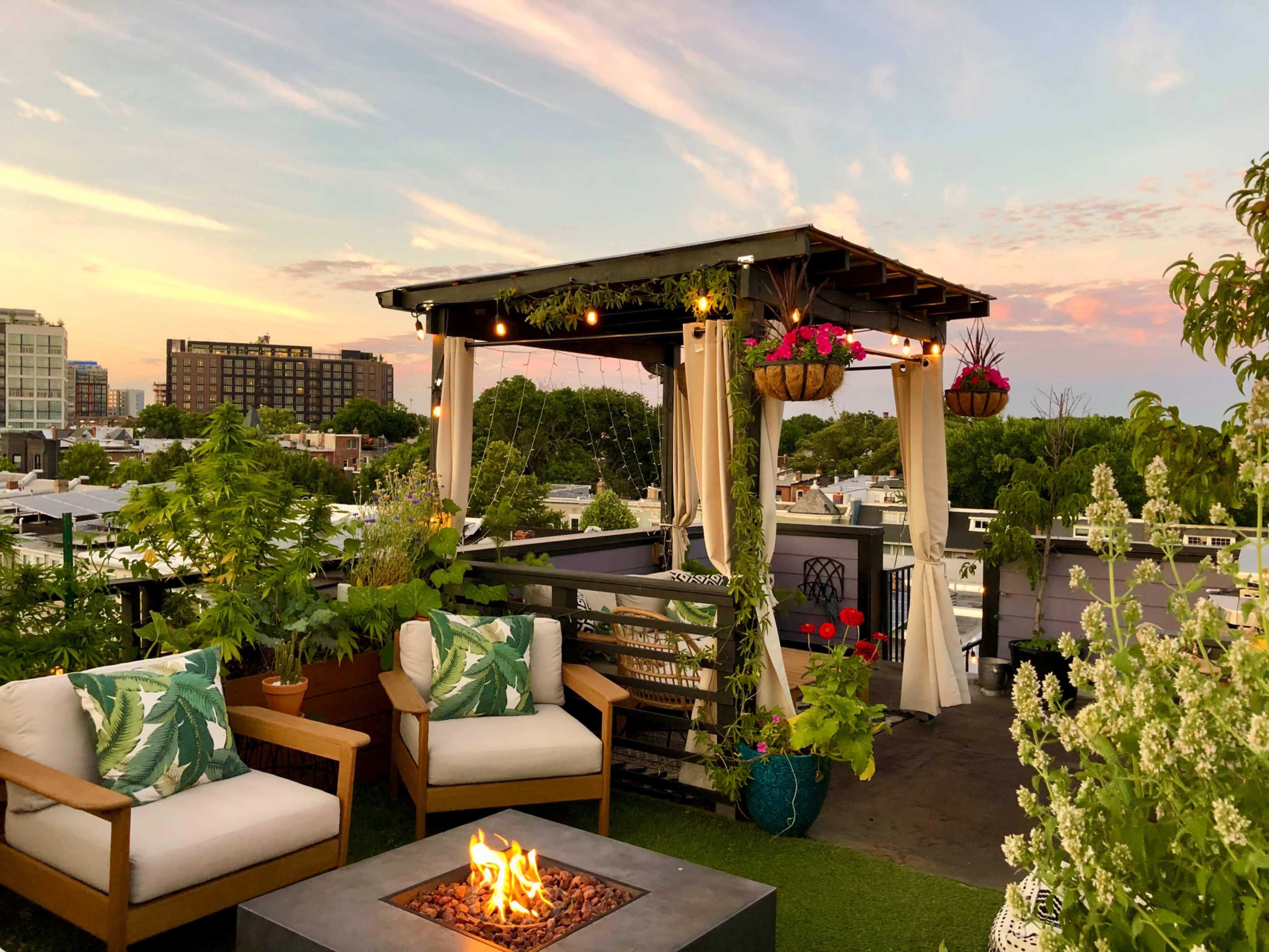A rooftop deck features a pergola adorned with hanging plants, cozy seating, and a fire pit, set against a colorful sunset.