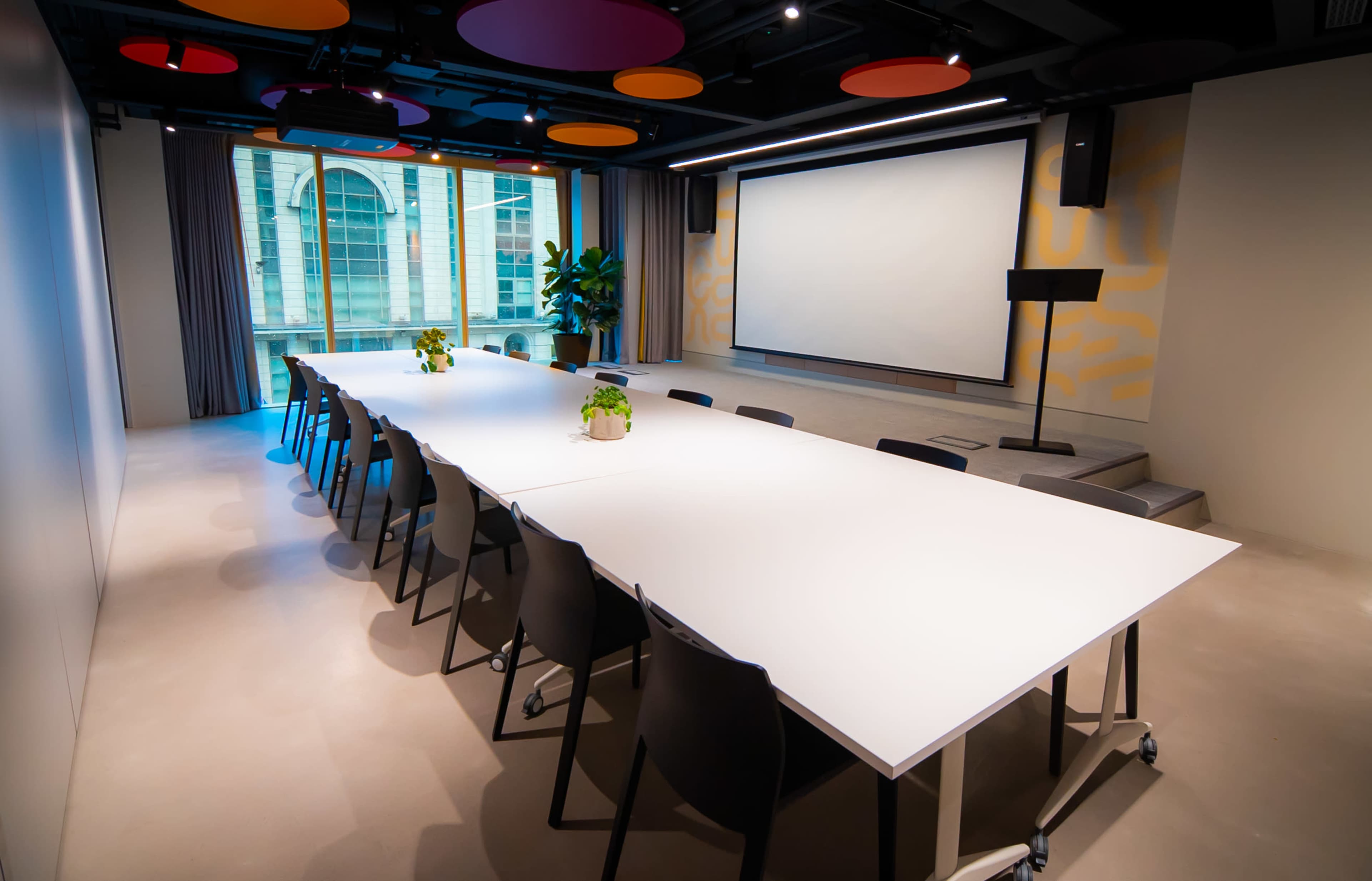 A modern conference room features a long white table with black chairs, a large screen at one end, and vibrant ceiling designs.