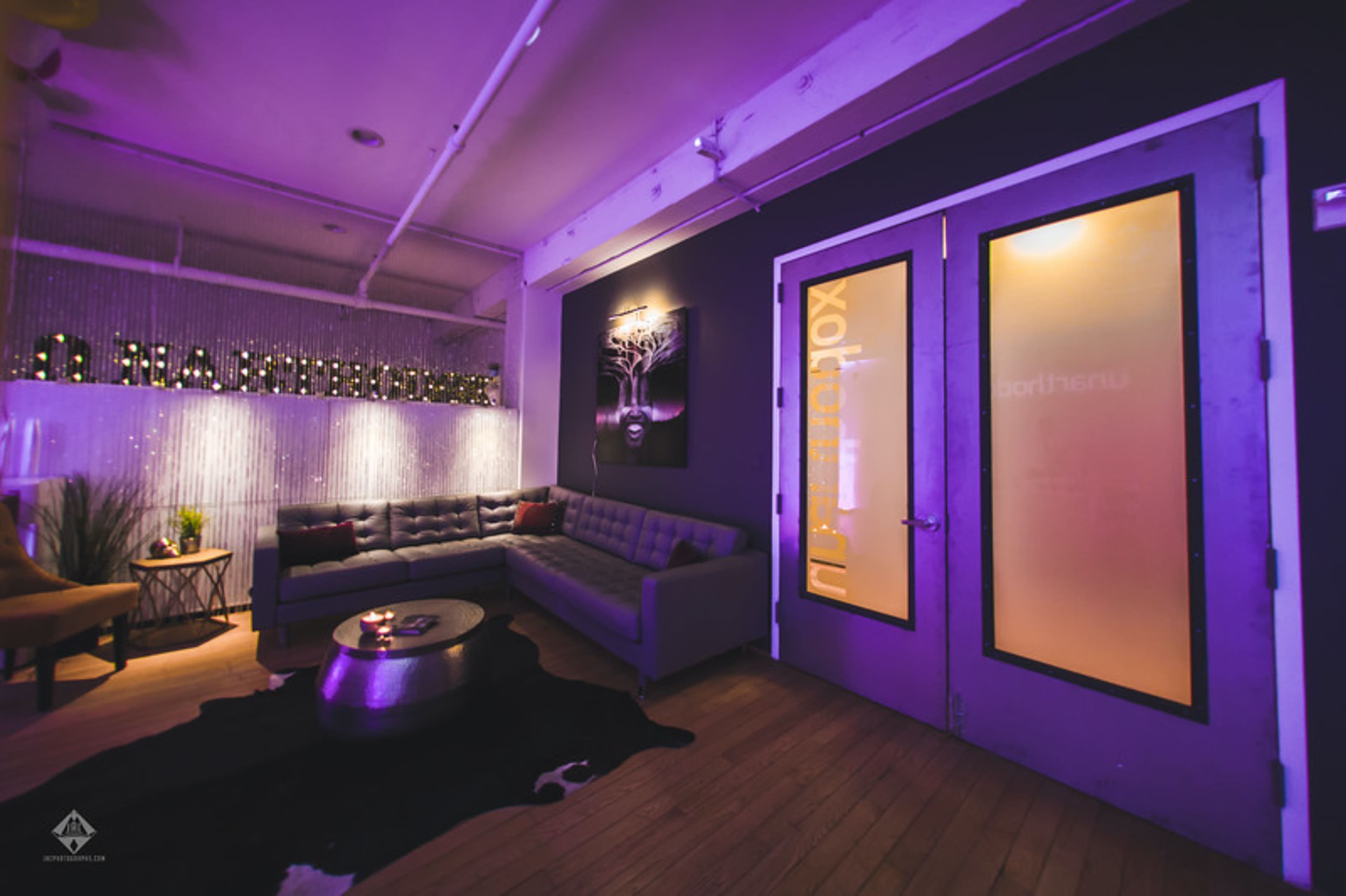 A modern interior with a purple glow featuring a gray sofa, a round table, and two glass doors against a dark wall.