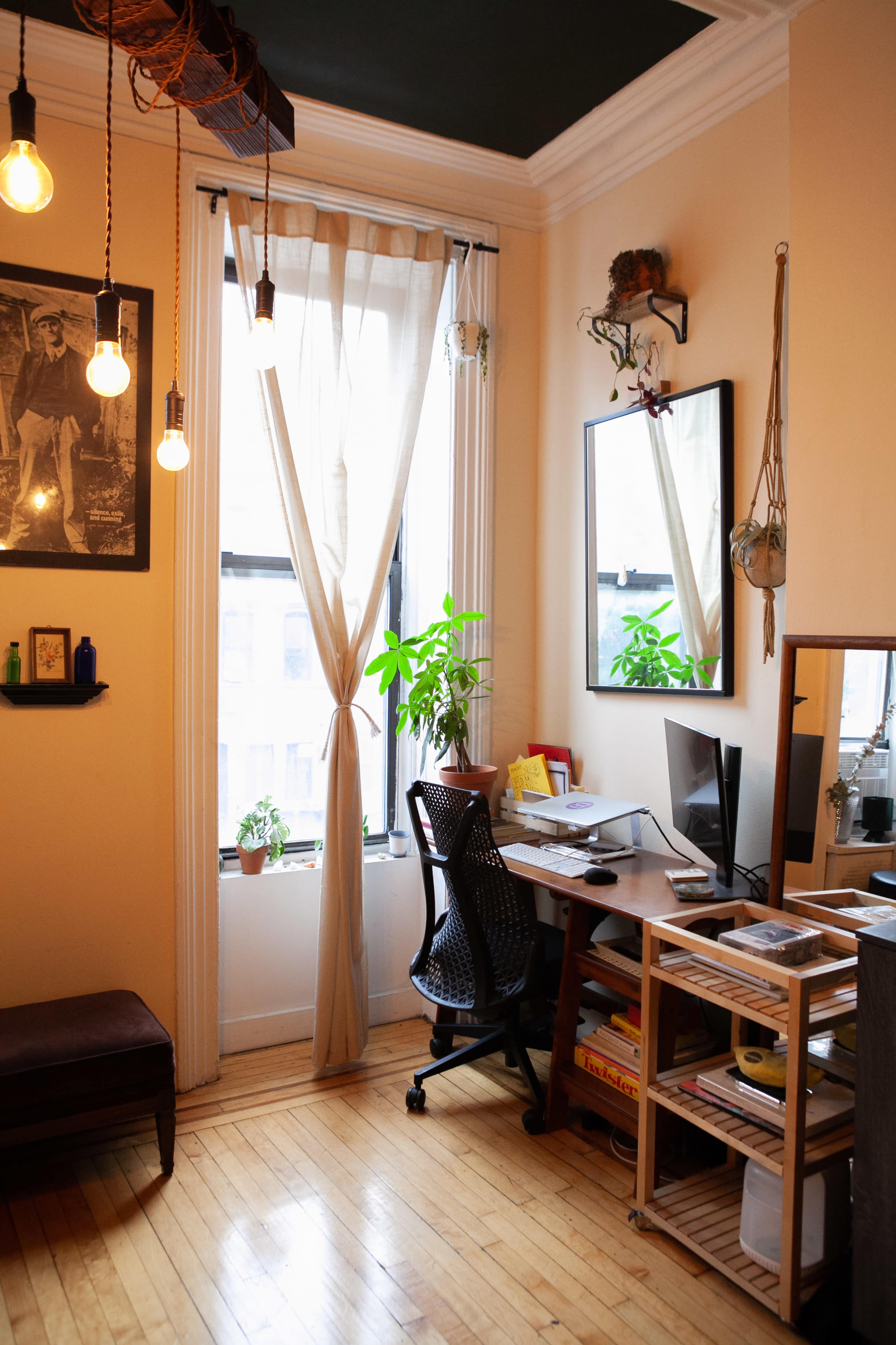 A small home office with a desk and computer, a chair, potted plants by the window, and light fixtures hanging from the ceiling.