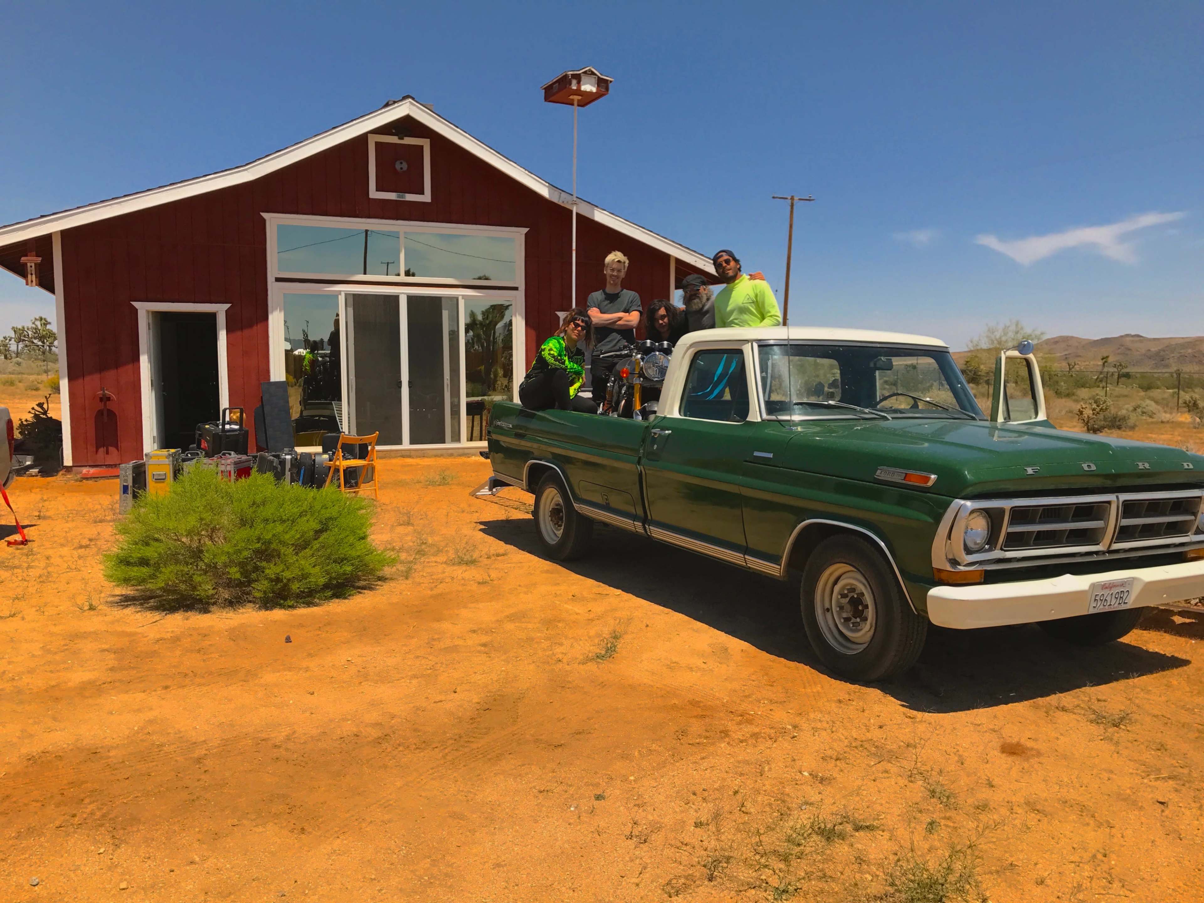 A group of four people is posed on a green truck parked in front of a red building in a desert landscape.