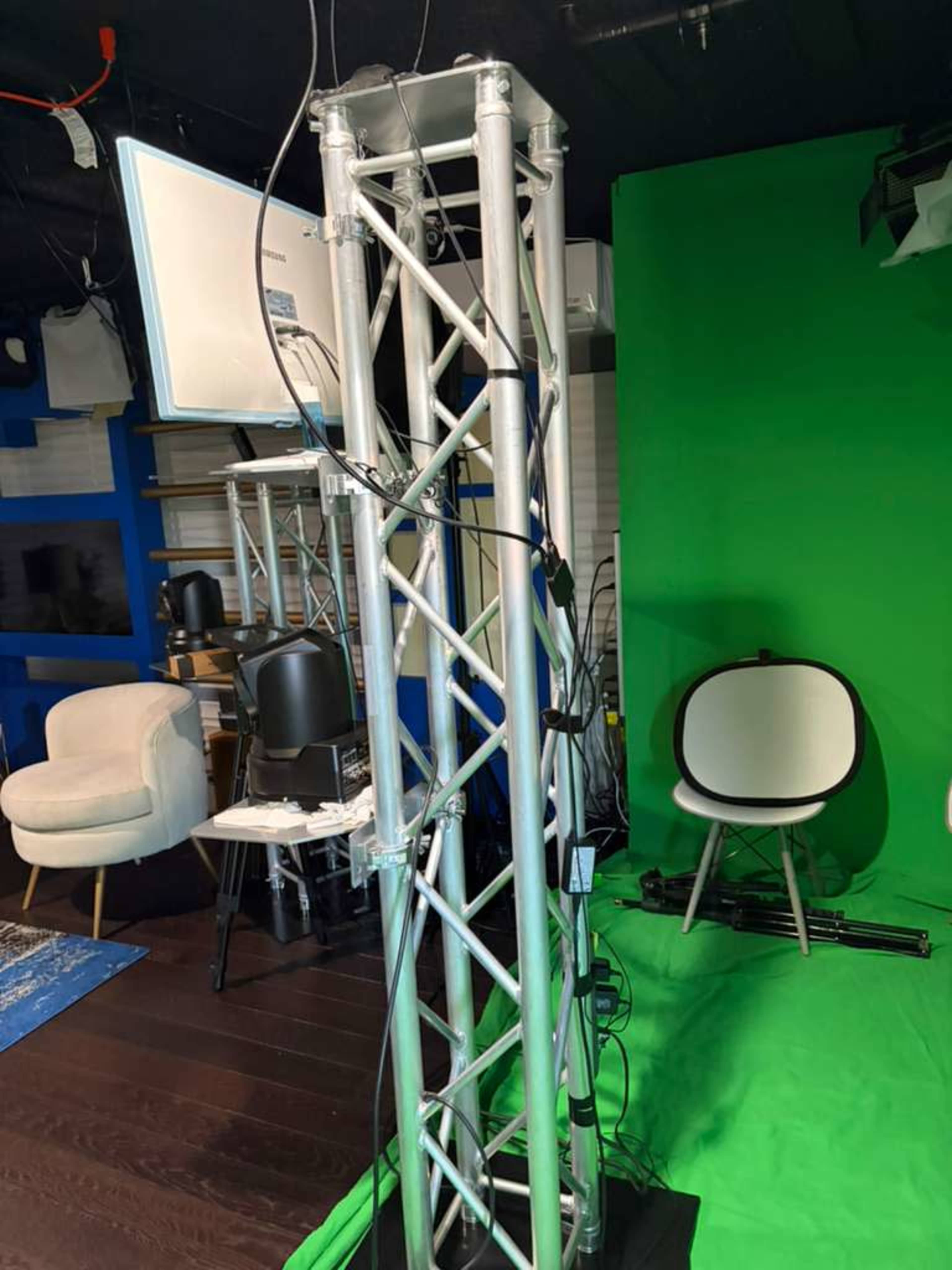 The image shows a metallic truss structure with cables and a monitor, situated in front of a green backdrop in a studio setting.