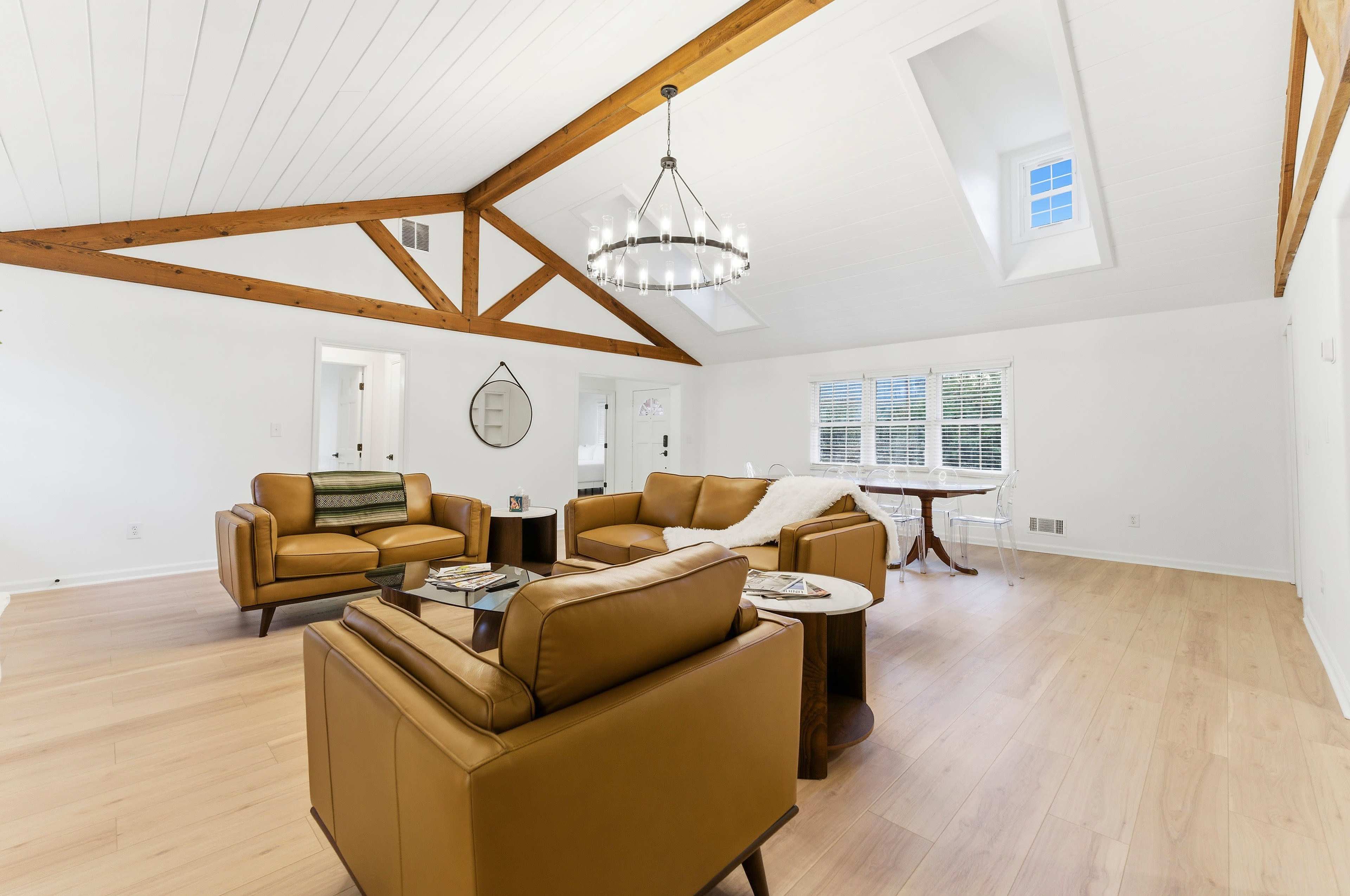 The image shows a well-lit living room featuring two leather sofas, a wooden dining table, and exposed wooden beams on the ceiling.