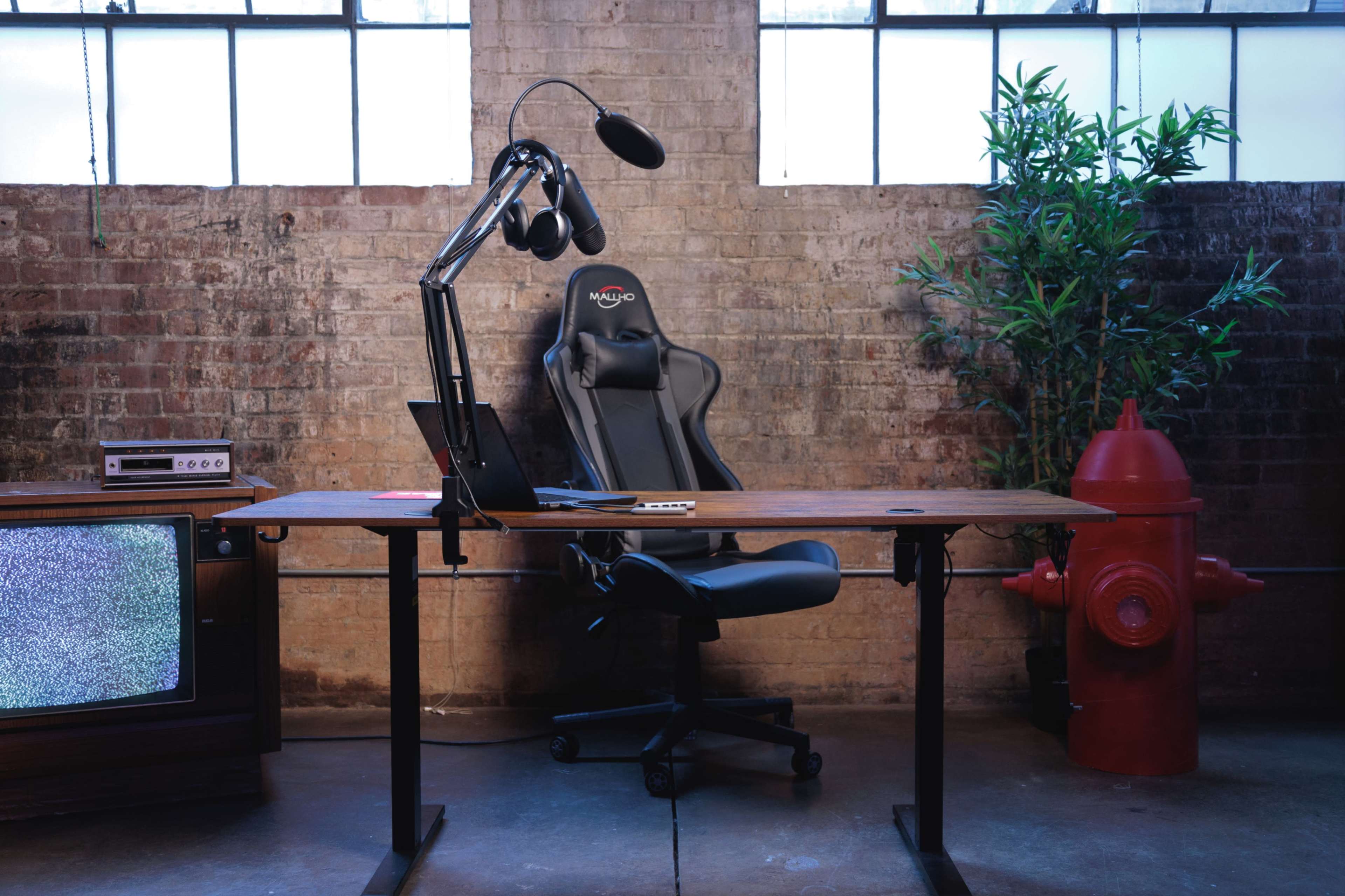 The image shows a modern workspace with an ergonomic black gaming chair, a wooden desk with a laptop, a microphone on an adjustable arm, a vintage television, and a red fire hydrant in an industrial setting with exposed brick walls.