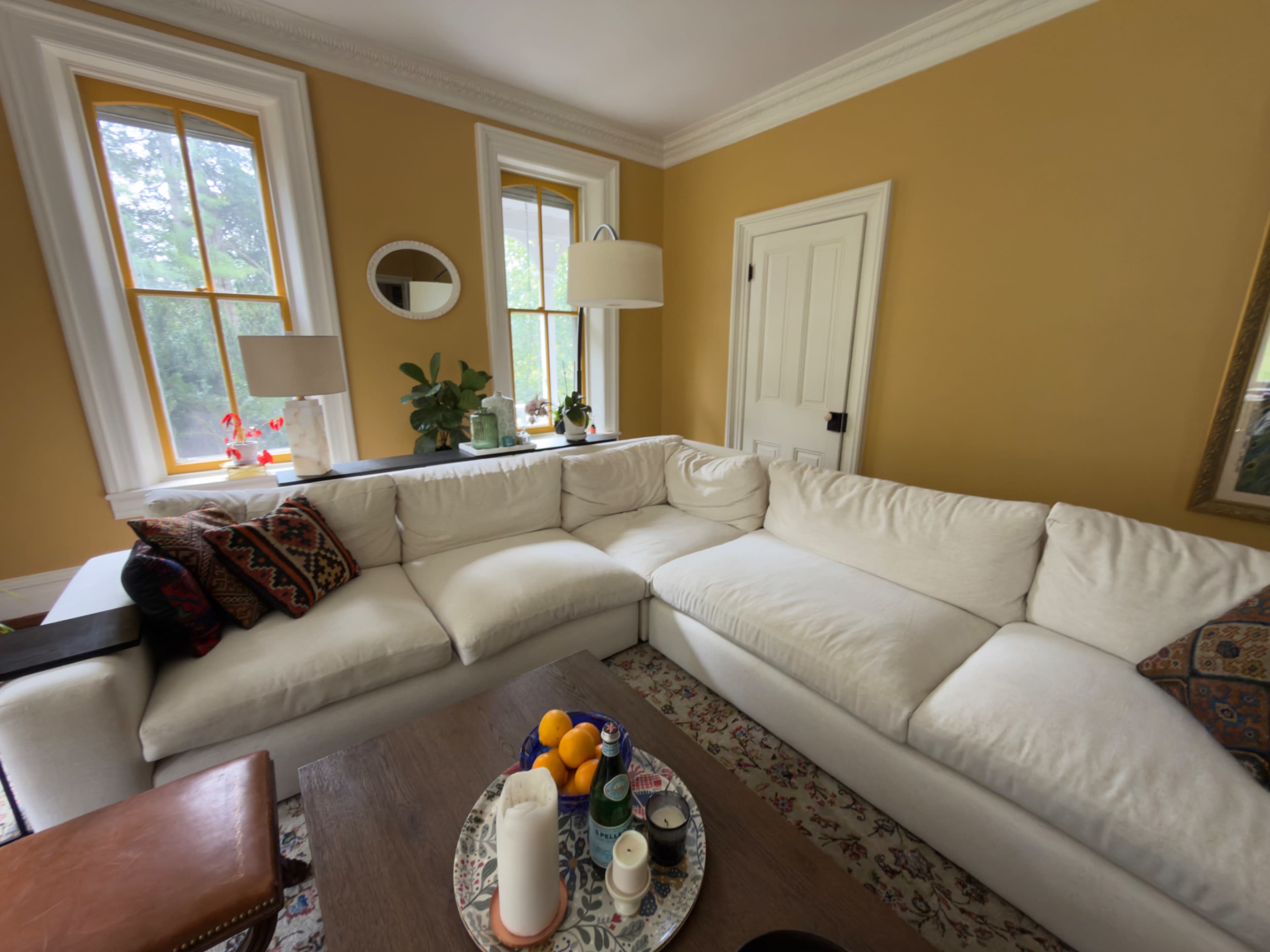The image shows a bright living room with a large white couch, a coffee table with fruits and drinks, and yellow walls adorned with windows and decorative items.