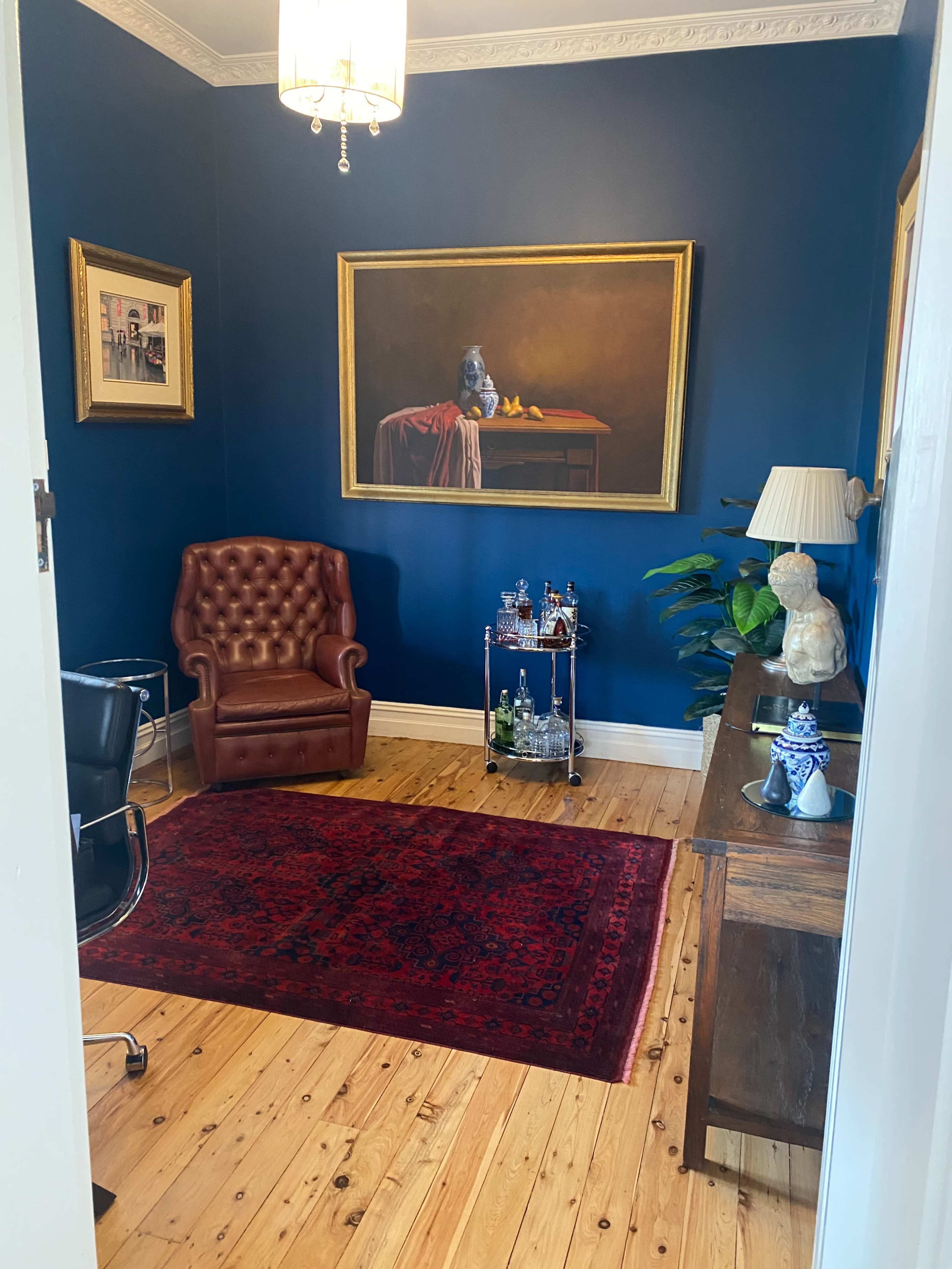 A cozy room with dark blue walls, featuring a brown leather armchair, a wooden side table with a lamp, a framed painting, and a small bar cart stocked with drinks.