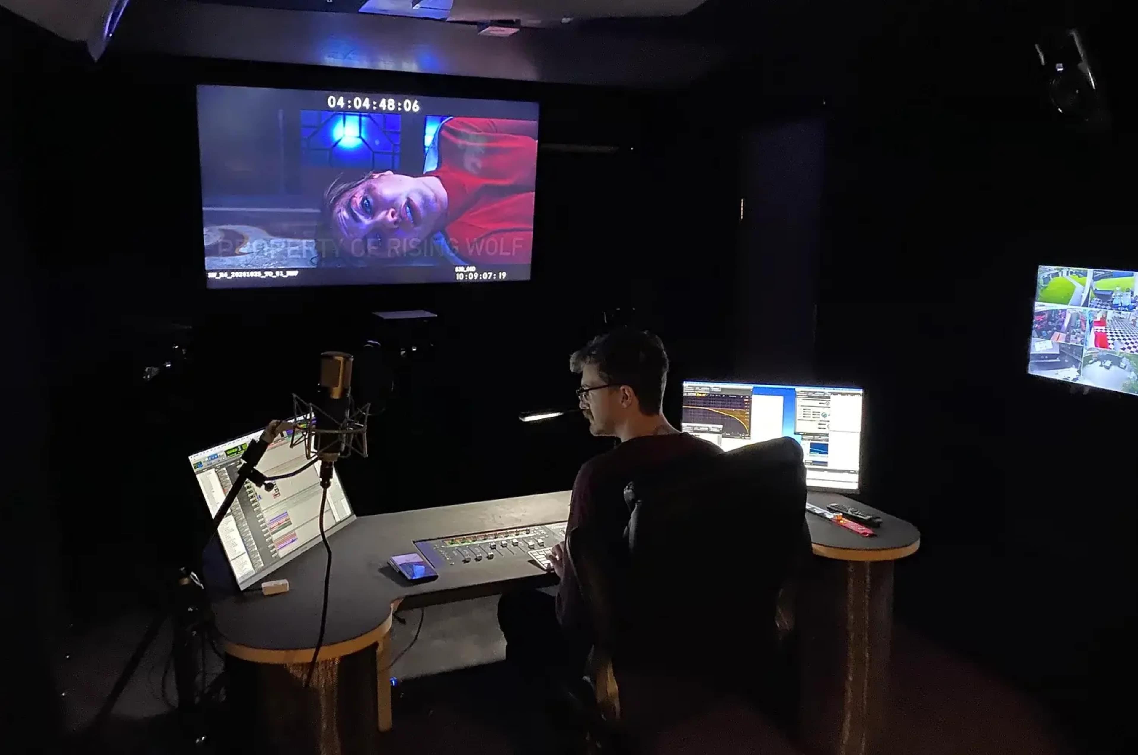 A person sits at a control desk in a dark studio, monitoring a screen displaying a scene from a film.