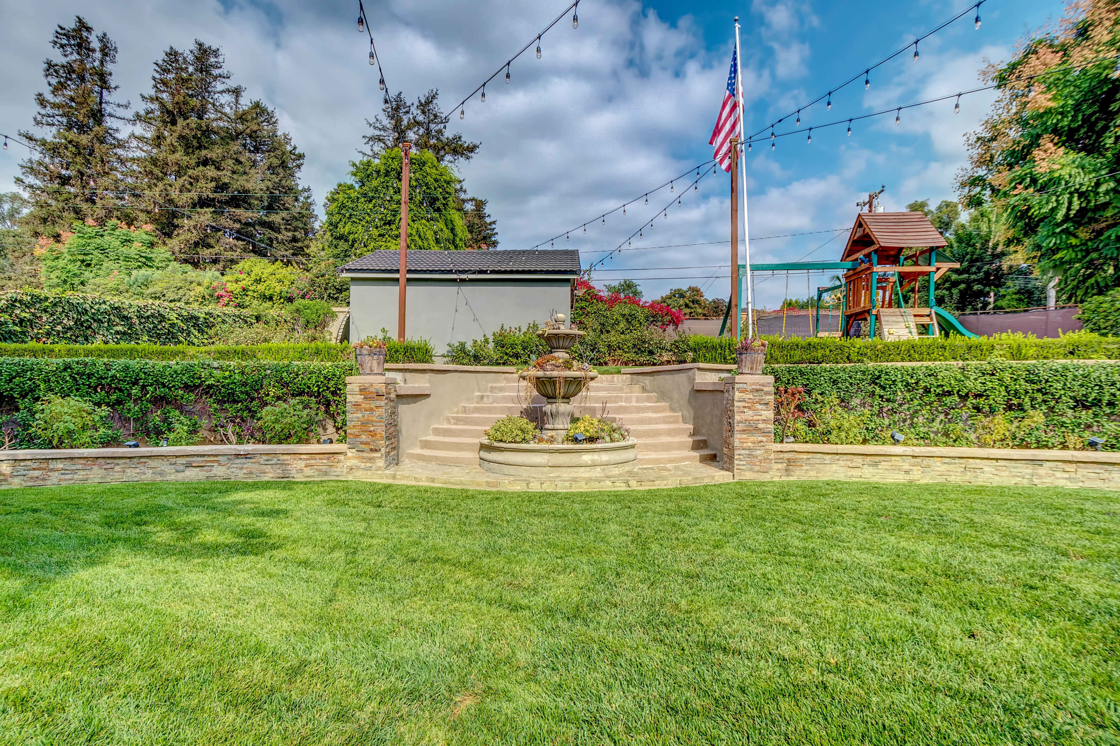 A stone fountain is centered in a landscaped yard, surrounded by neatly trimmed hedges and a playset in the background under a cloudy sky.
