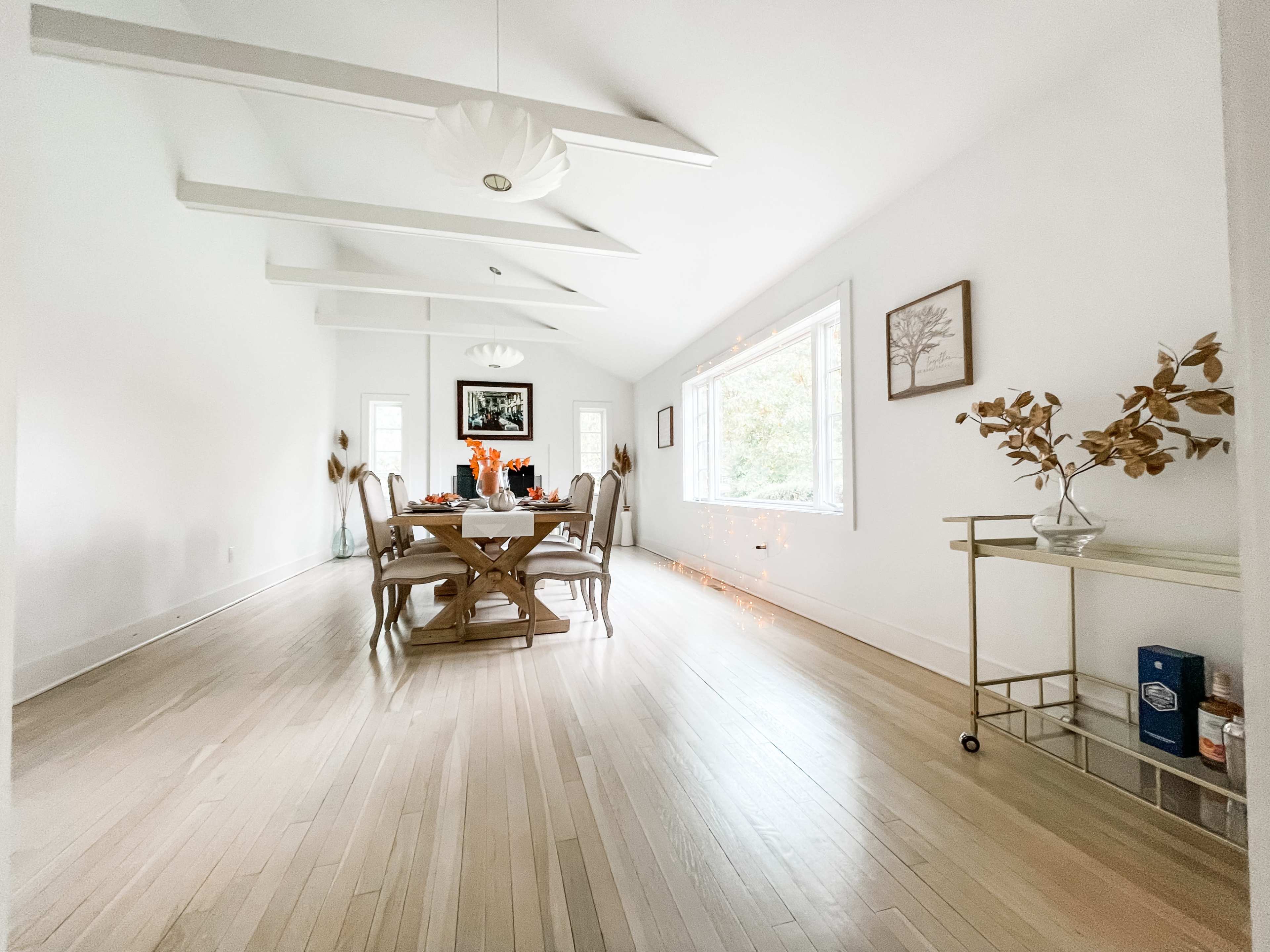 A dining area with a round table set for a meal, surrounded by light-colored walls and wooden floorboards, features large windows and decorative elements.