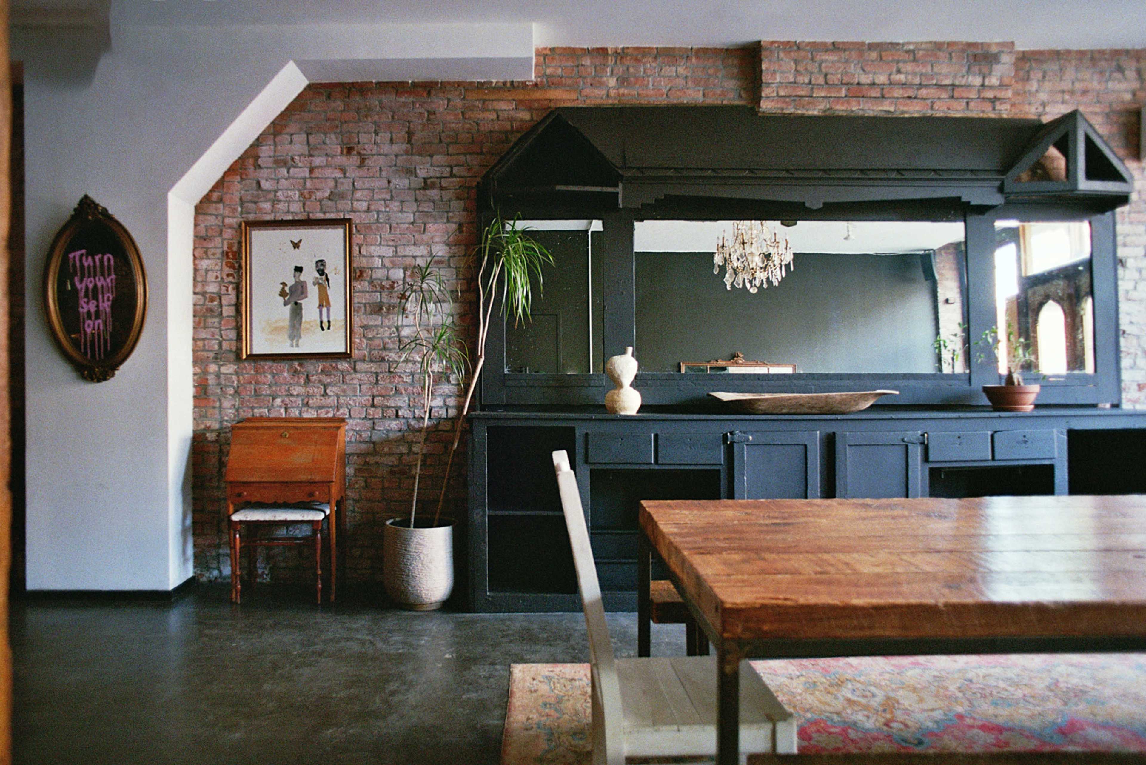 The image shows a dining area with a wooden table, black cabinetry, a decorative mirror, and exposed brick walls.