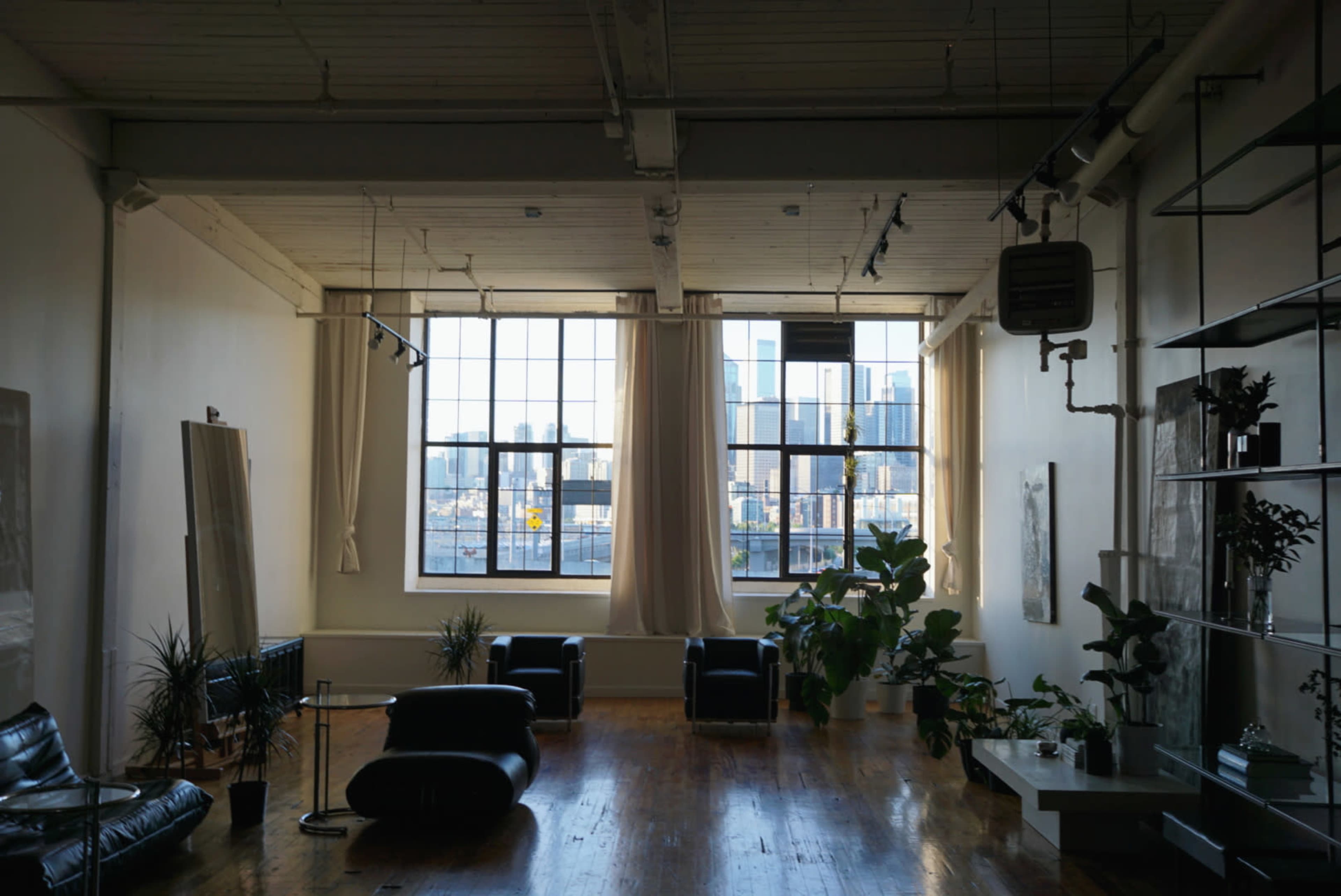 The image shows a spacious, modern loft with large windows, plants, and minimalist furniture that offers a view of a city skyline.
