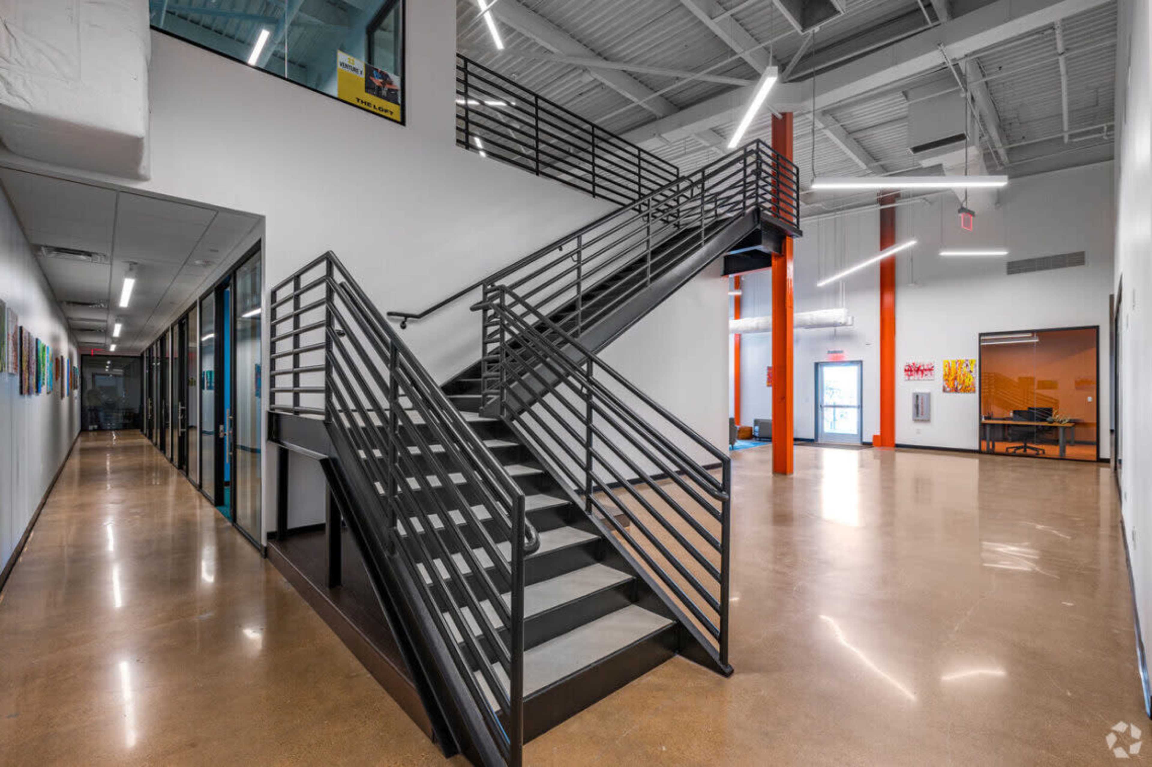 The image shows a modern interior space featuring a metal staircase, polished concrete floors, and colorful artwork along the walls.