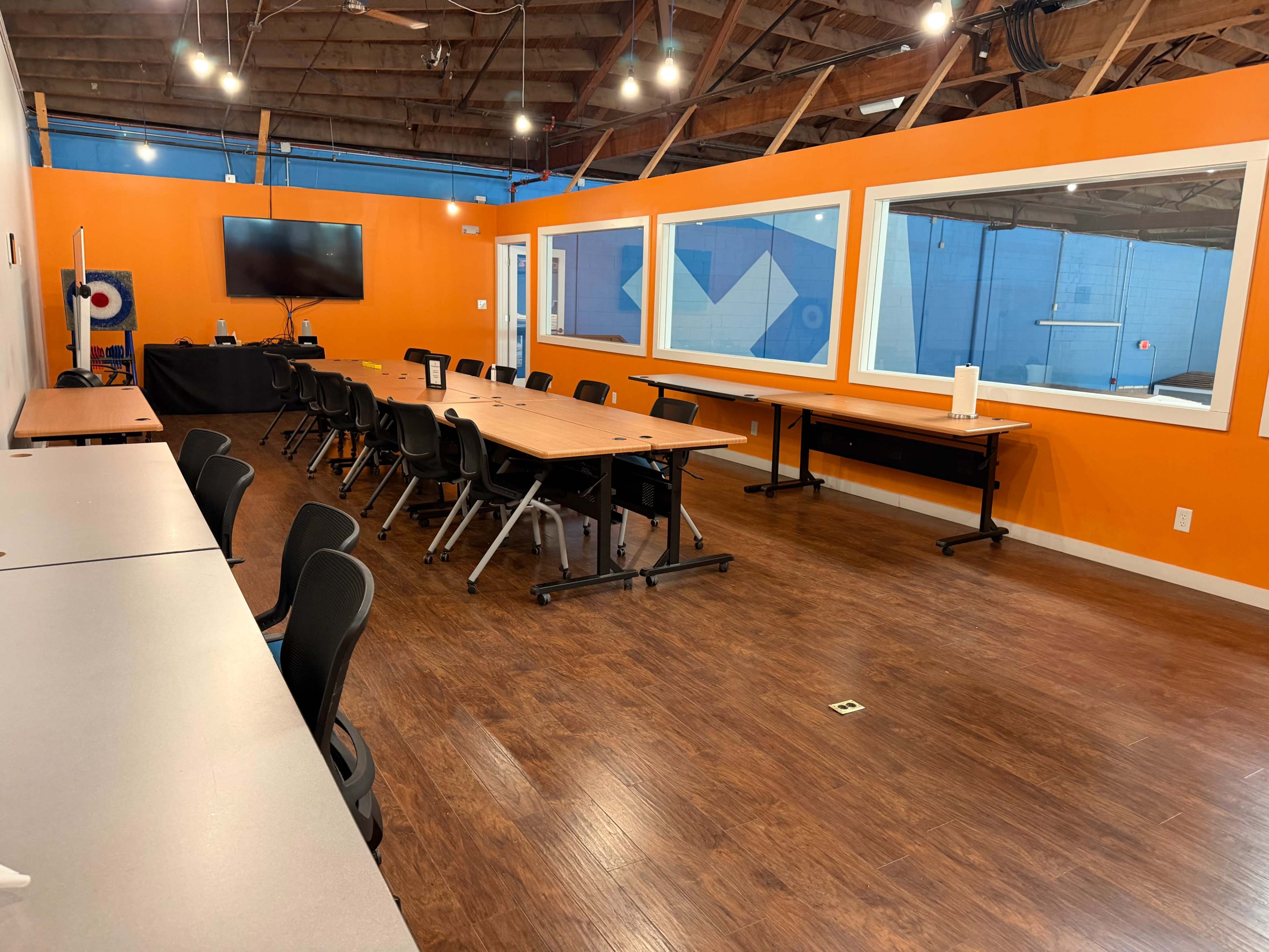 The image shows a spacious meeting room with multiple tables and chairs arranged for collaboration, featuring an orange wall and a television mounted on one side.