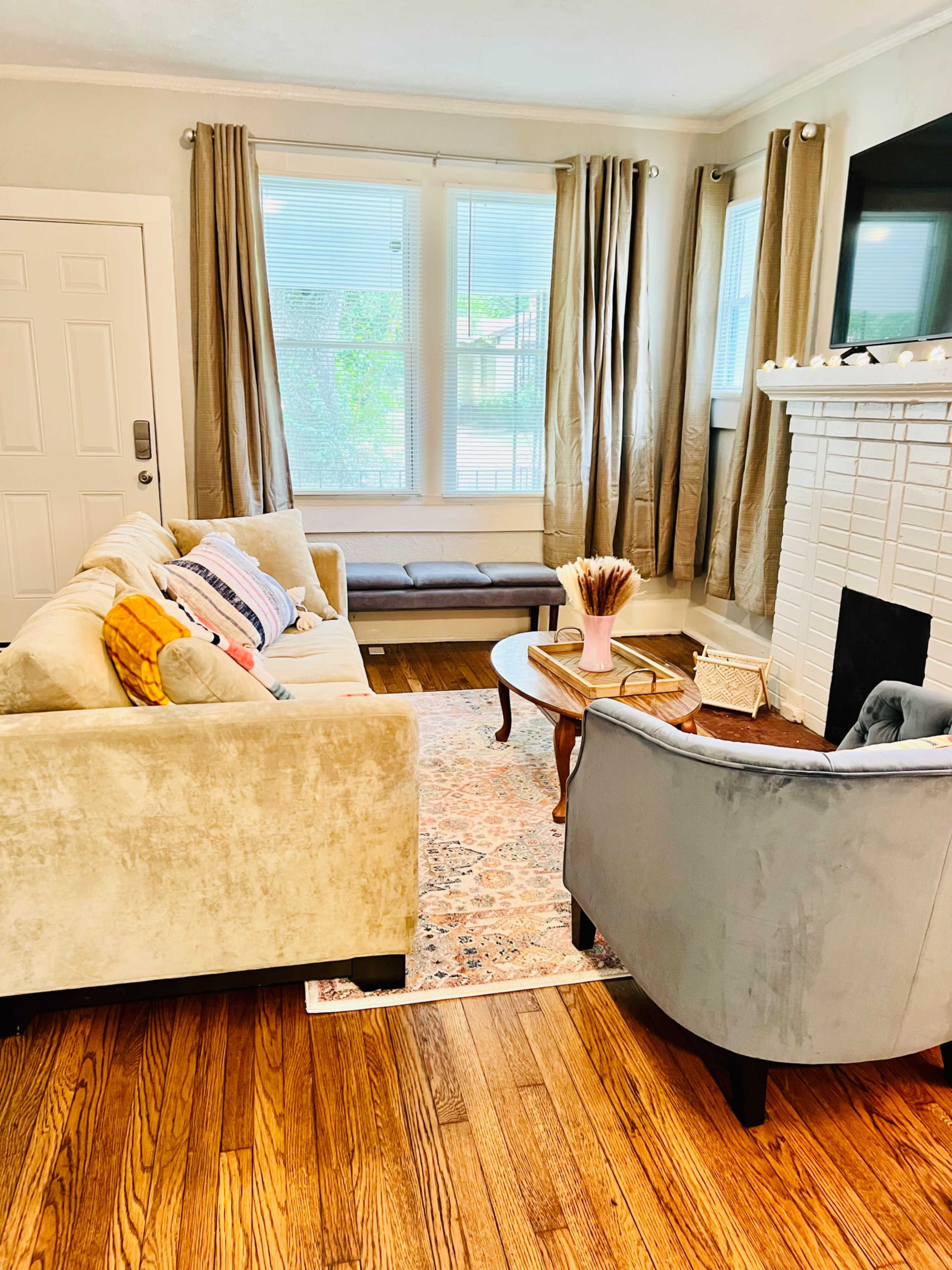 A cozy living room features a light-colored sofa, a gray armchair, a wooden coffee table, and a fireplace, with large windows allowing natural light to enter.