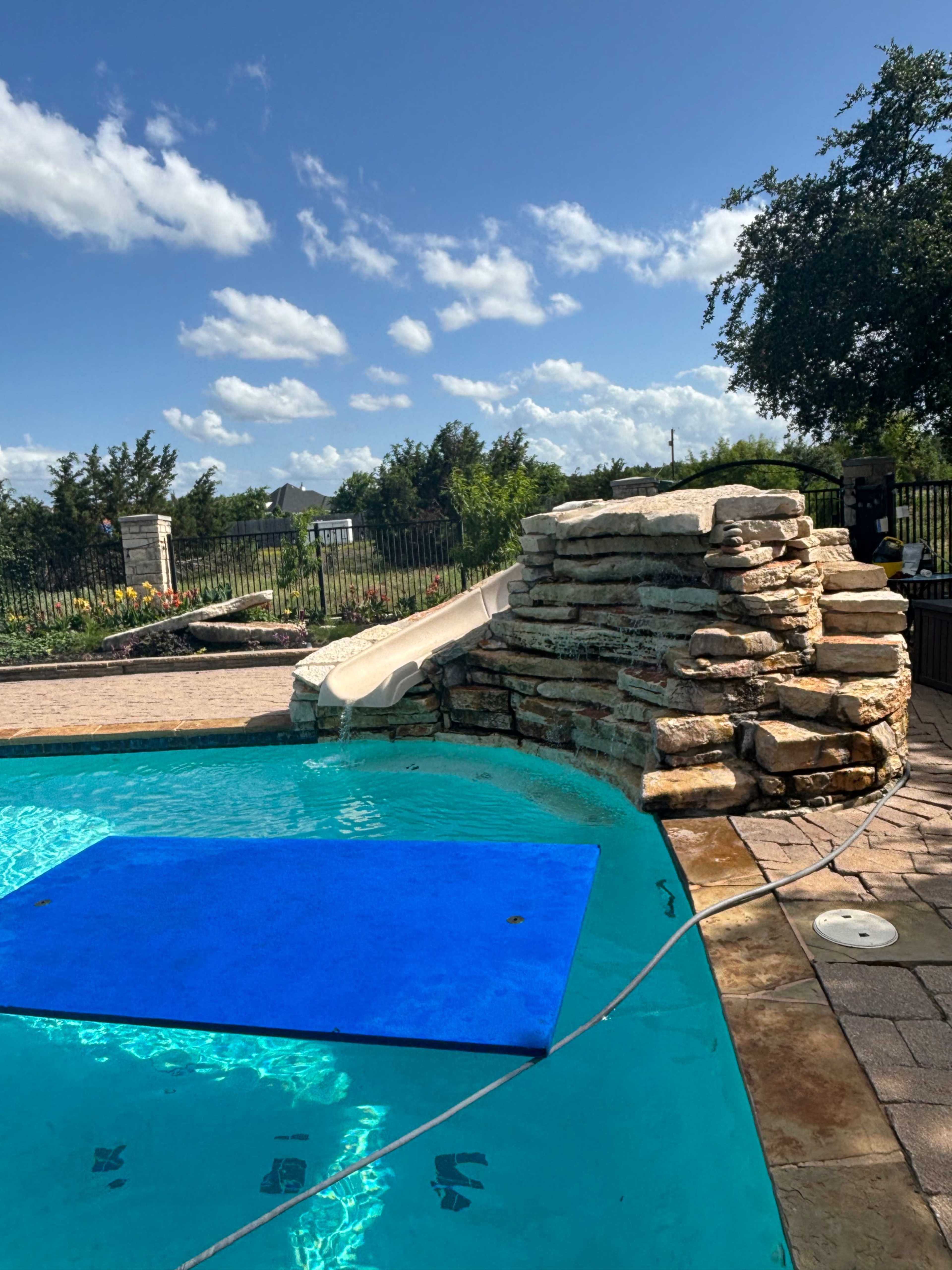 A pool with a blue cover over part of the water features a stone slide and a surrounding landscaped yard under a partly cloudy sky.