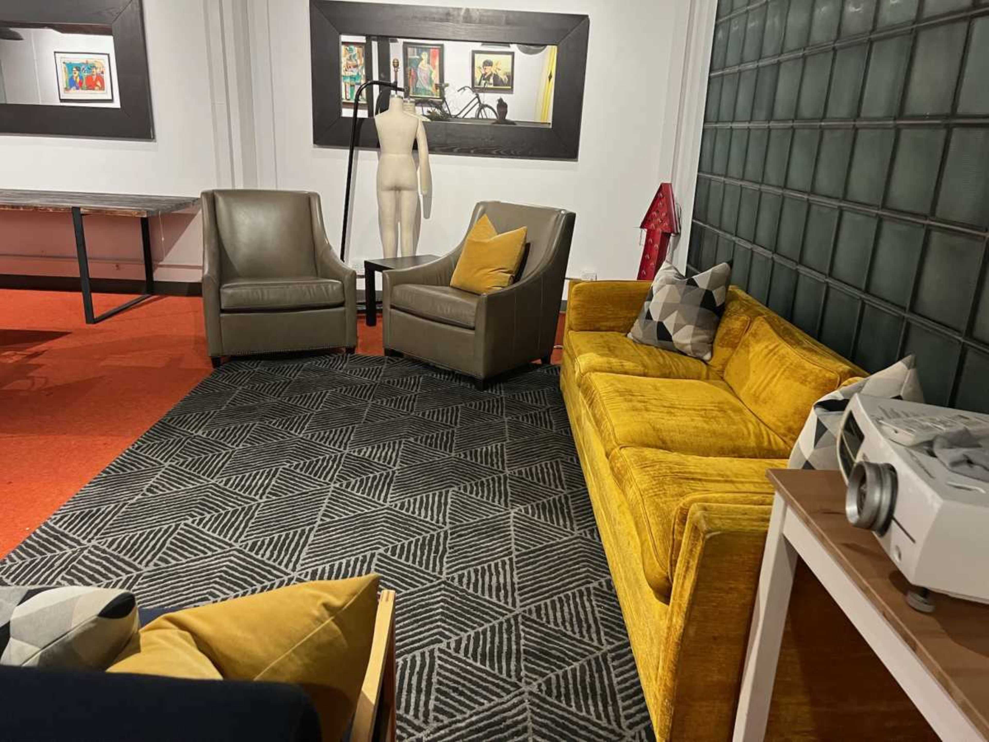 The image shows a cozy seating area featuring a yellow couch, two gray armchairs, a geometric patterned rug, and a backdrop of framed artwork and a mannequin.