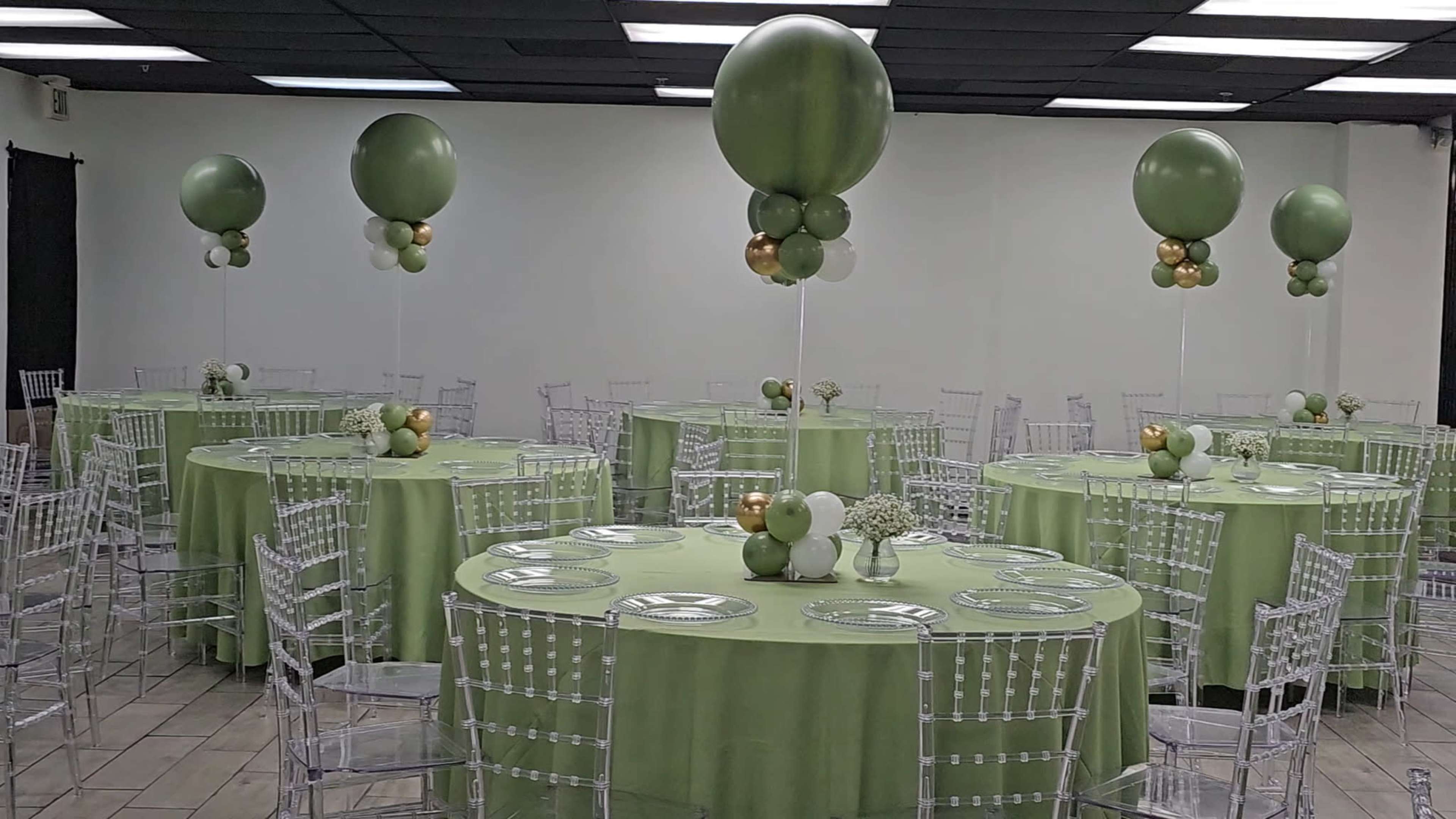 The image shows a banquet hall set up with several round tables covered in light green tablecloths, each adorned with balloon centerpieces and clear chiavari chairs.