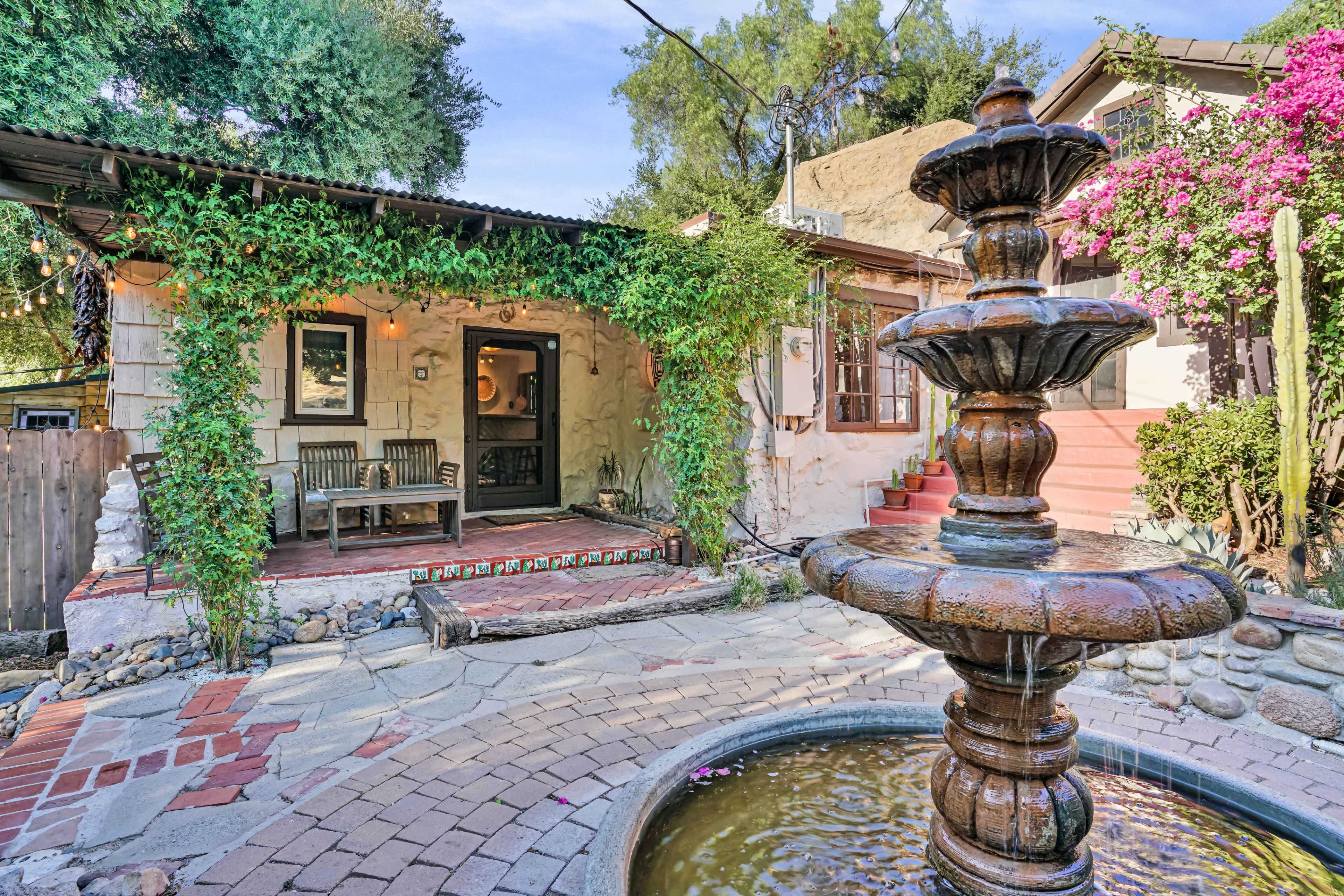 The image shows a courtyard with a stone fountain in the foreground, surrounded by a patio area featuring a wooden bench and lush greenery.