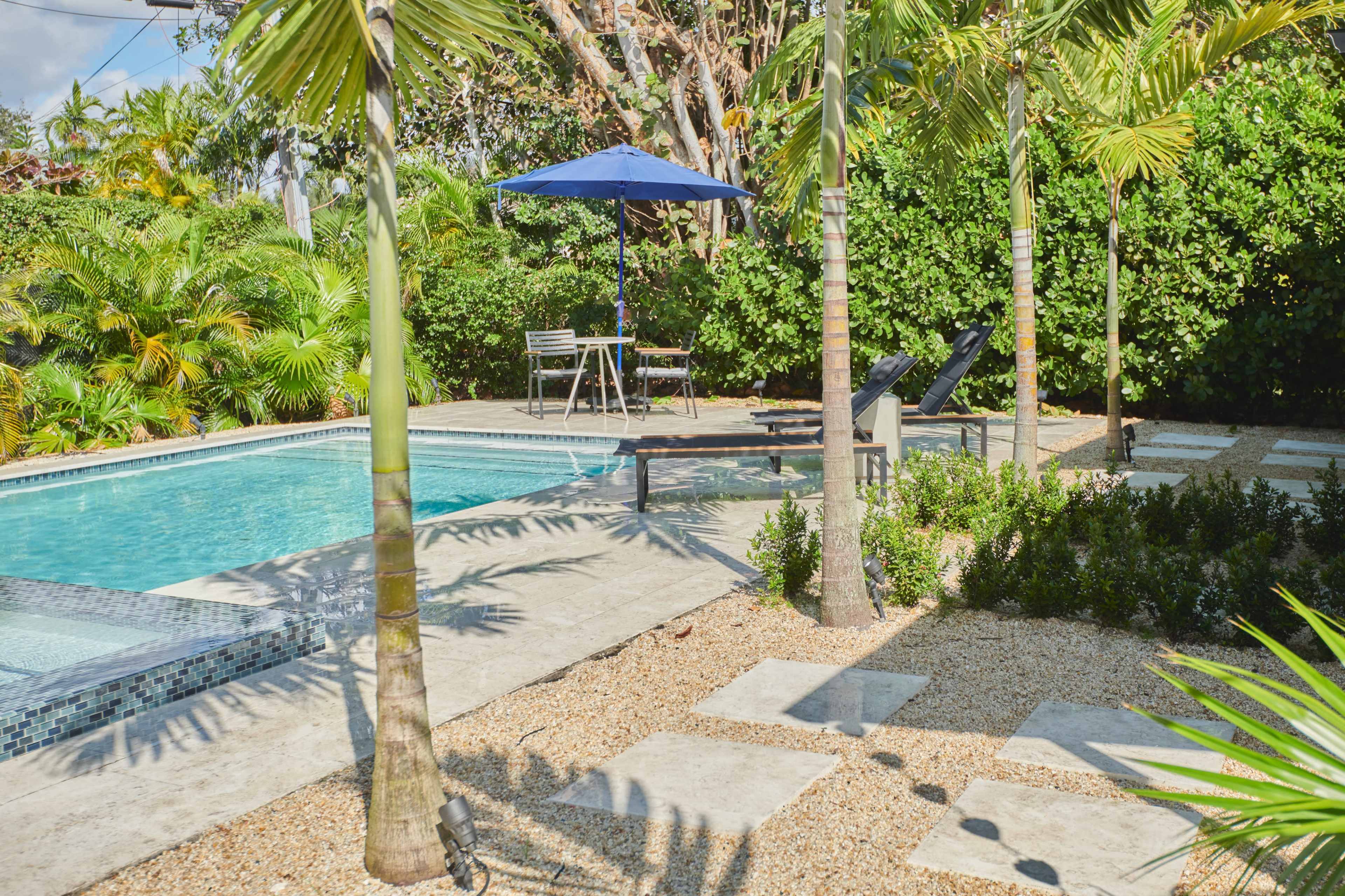 A swimming pool surrounded by palm trees, lounge chairs, and a small dining area with a table and umbrella.