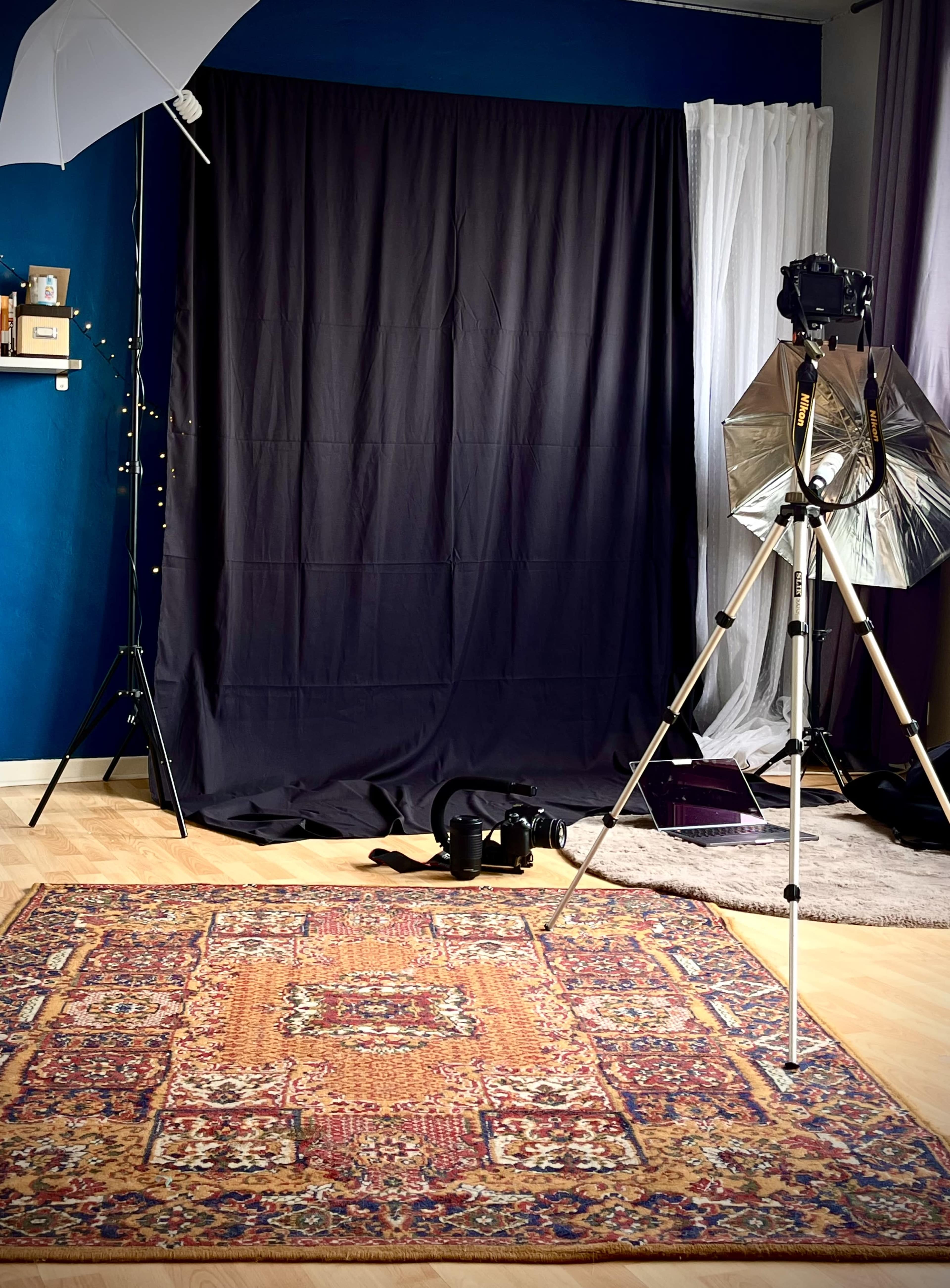 The image shows a photography studio setup with a black backdrop, a patterned rug on the floor, lighting equipment, and a camera on a tripod.