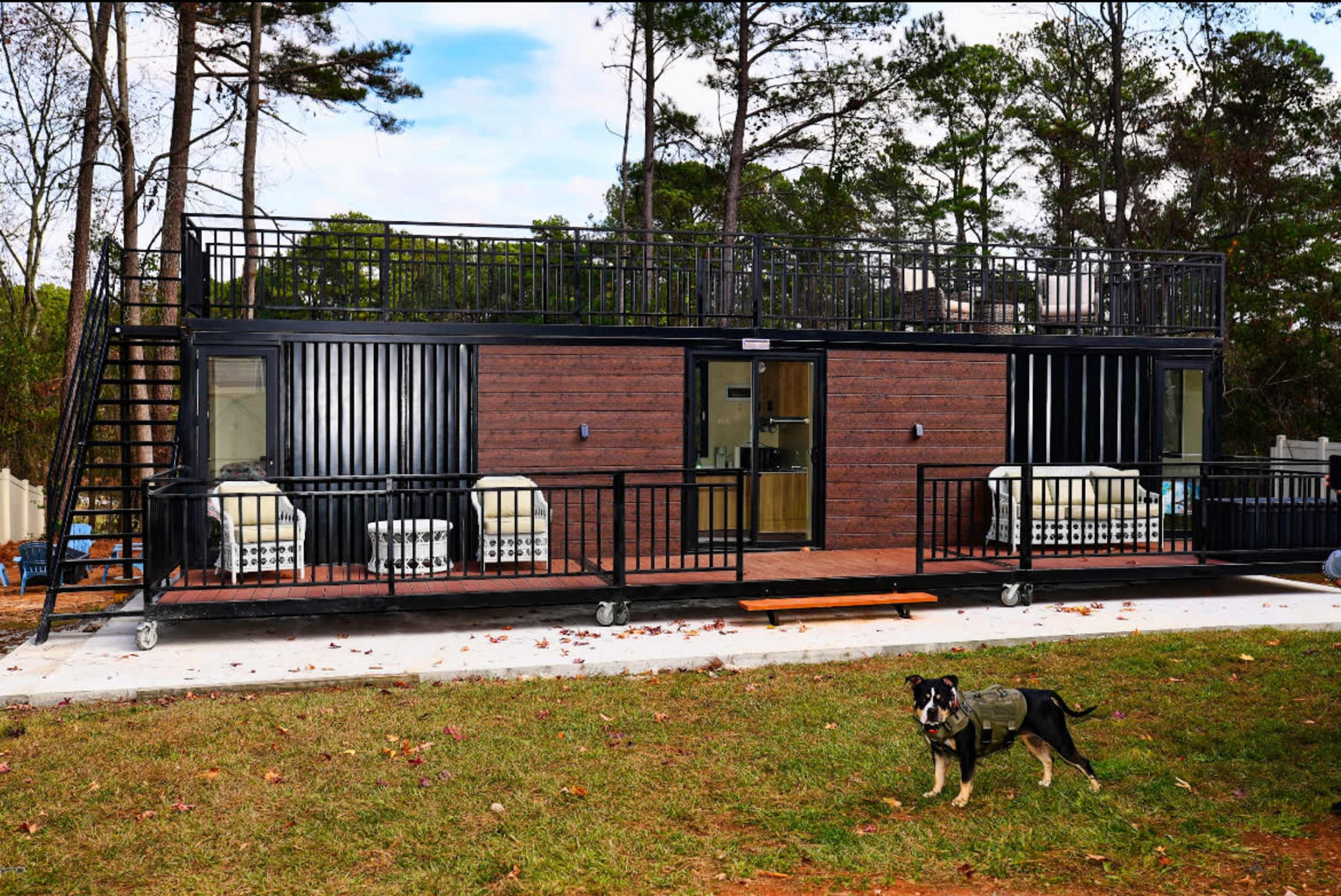 A modern, two-story shipping container home features a deck and seating area in front, with a dog in the foreground.