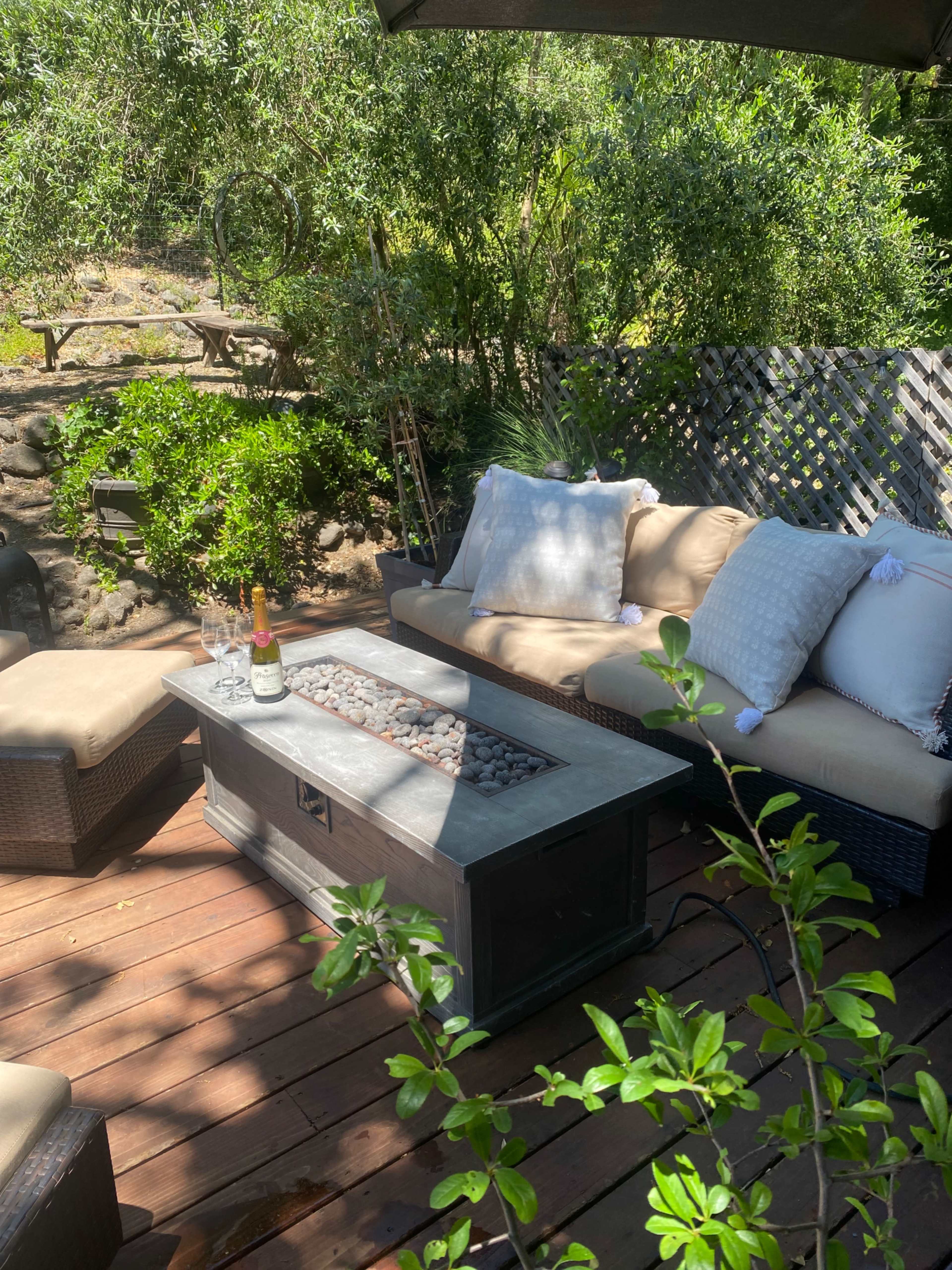 A patio area features a sectional couch with plush pillows, a stone fire table, and two glasses beside a bottle of champagne, surrounded by greenery.