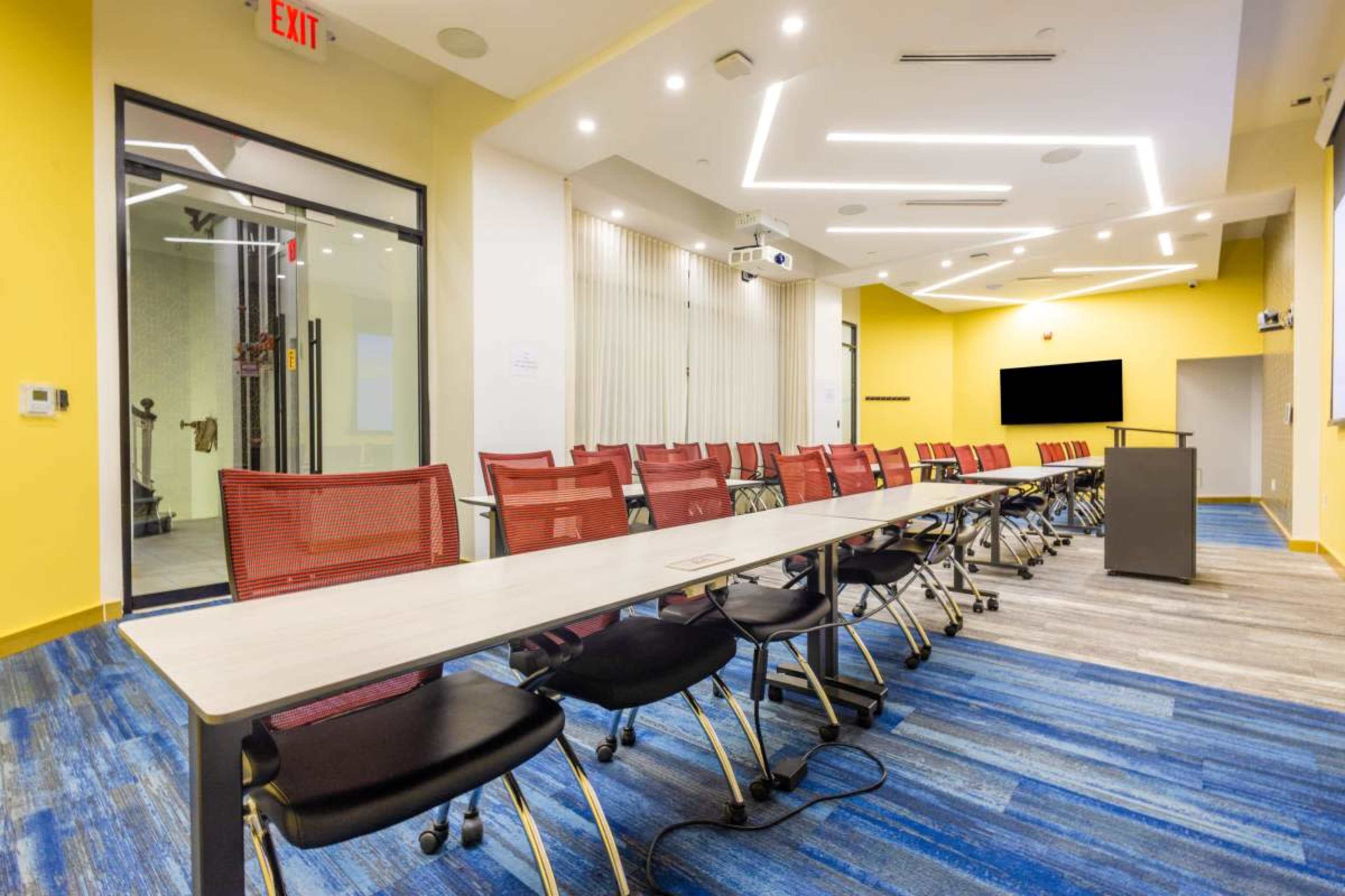 The image shows a modern conference room with a long table lined with red chairs, featuring a projector and a large screen at one end.
