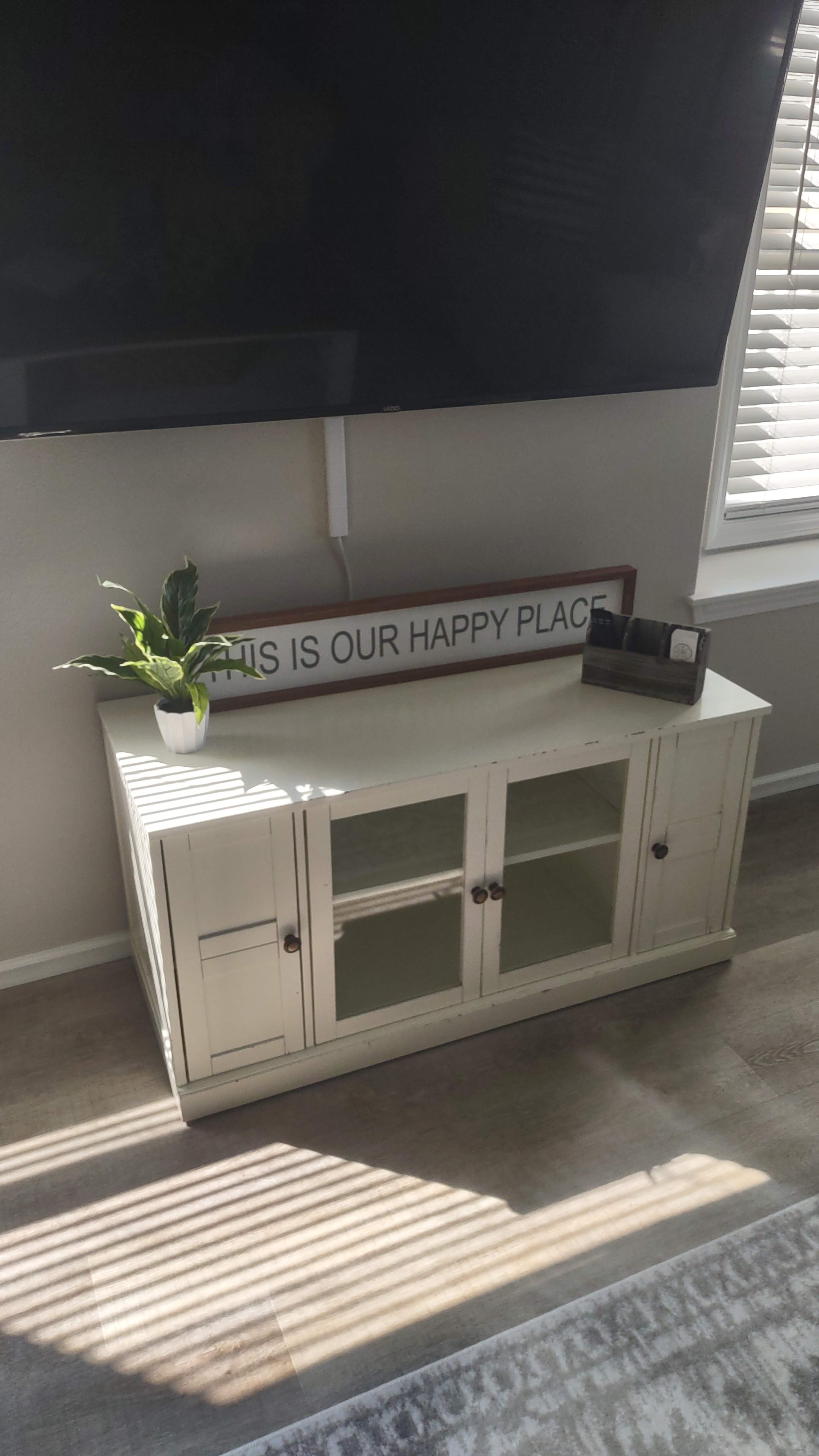 A white wooden TV stand with glass doors sits against a wall beneath a large screen, featuring a sign that reads "THIS IS OUR HAPPY PLACE" and a small potted plant.