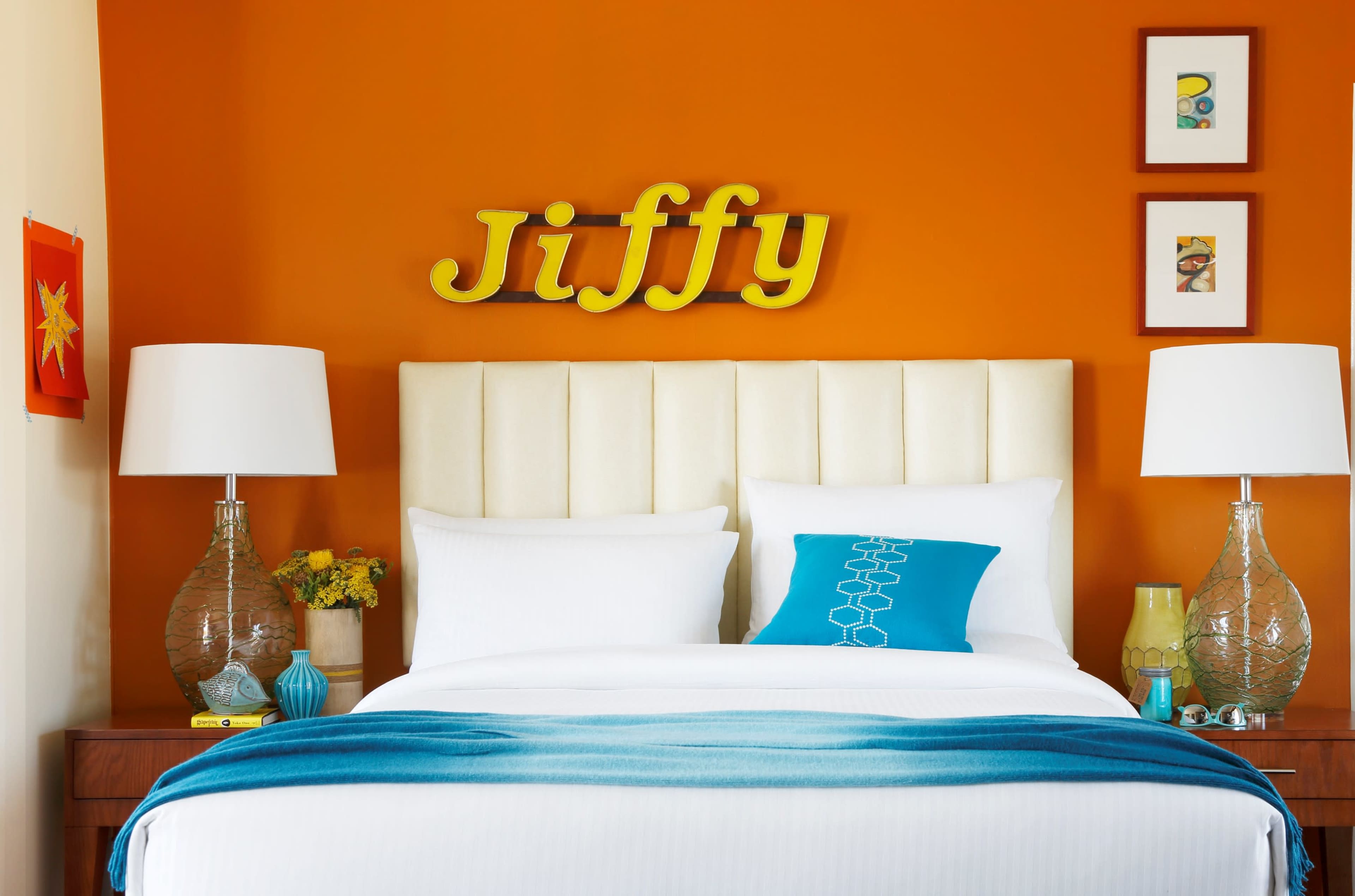 A bed with white linens and a blue accent pillow is placed against an orange wall featuring the word "Jiffy" in yellow and framed artwork.