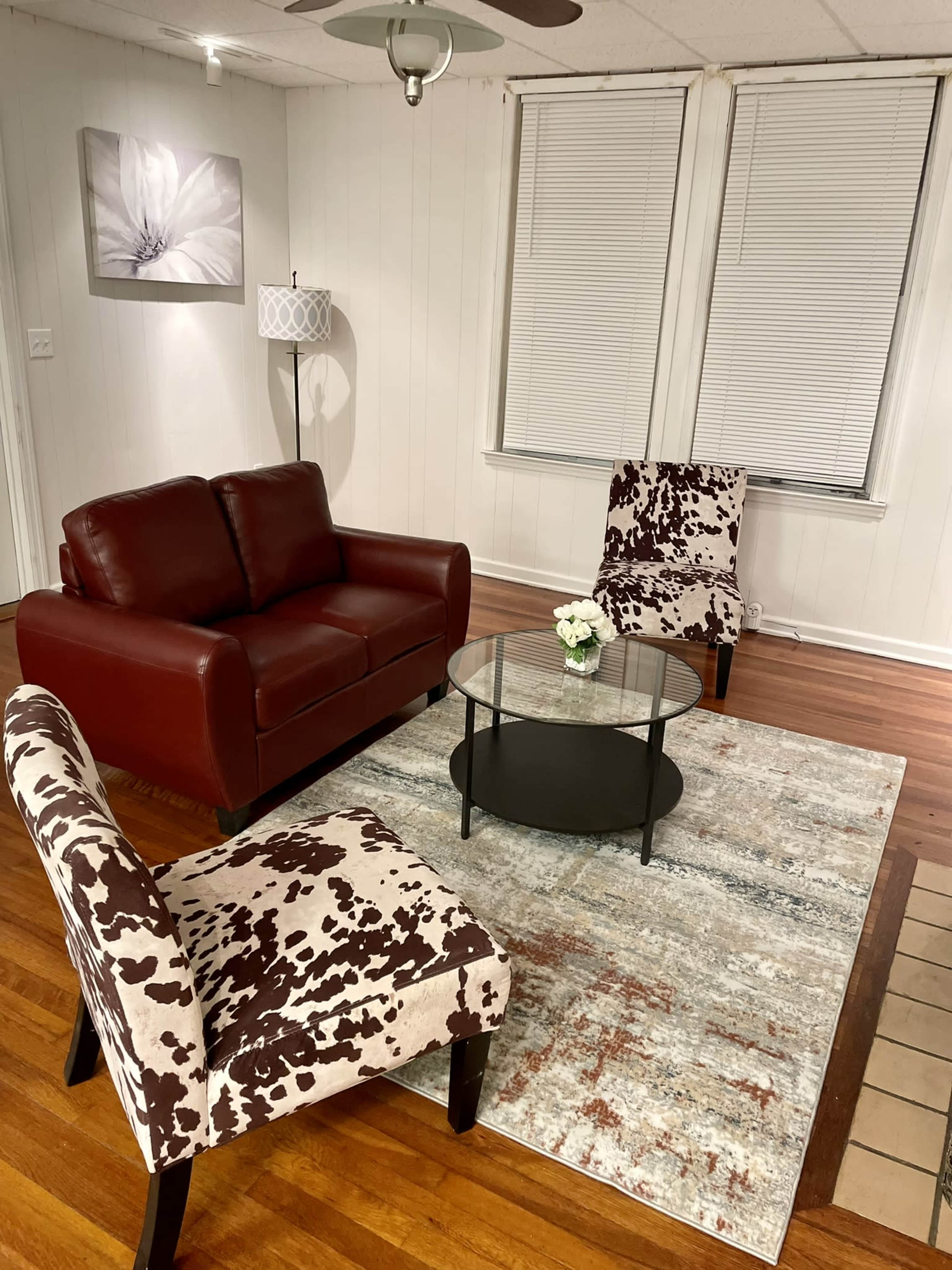 The image shows a living room with a red leather sofa, two patterned chairs, a glass coffee table on a rug, and a floral wall art piece.