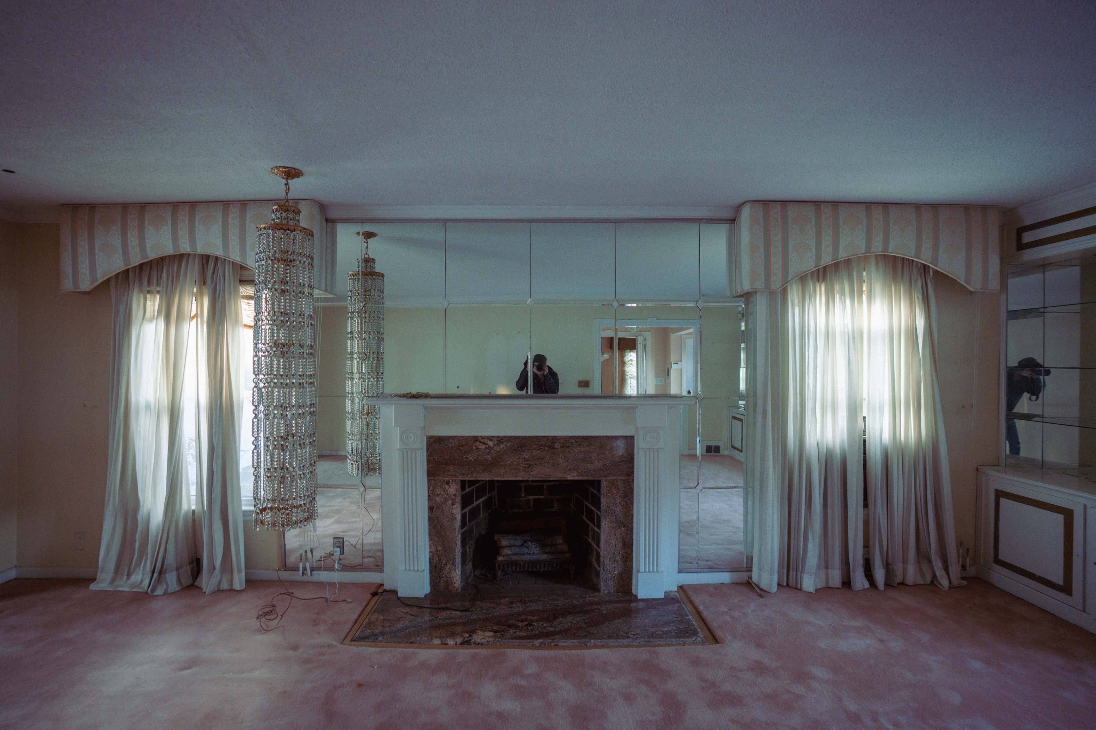 The image shows a living room with a fireplace, mirrored walls, two hanging chandeliers, and large curtains on the windows.