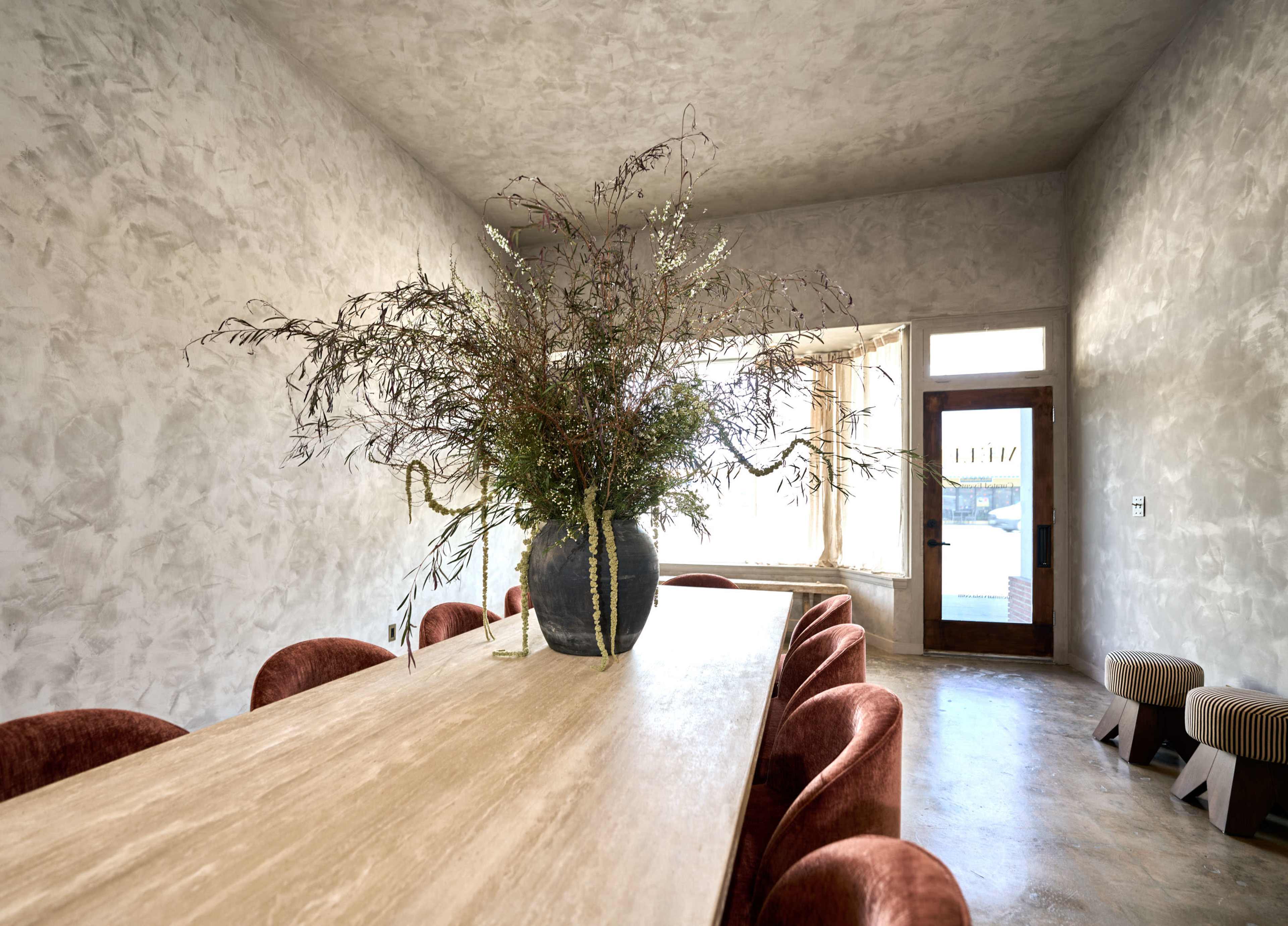 A long wooden table with eight upholstered chairs is set in a minimalistic room adorned with a large vase of greenery on top.