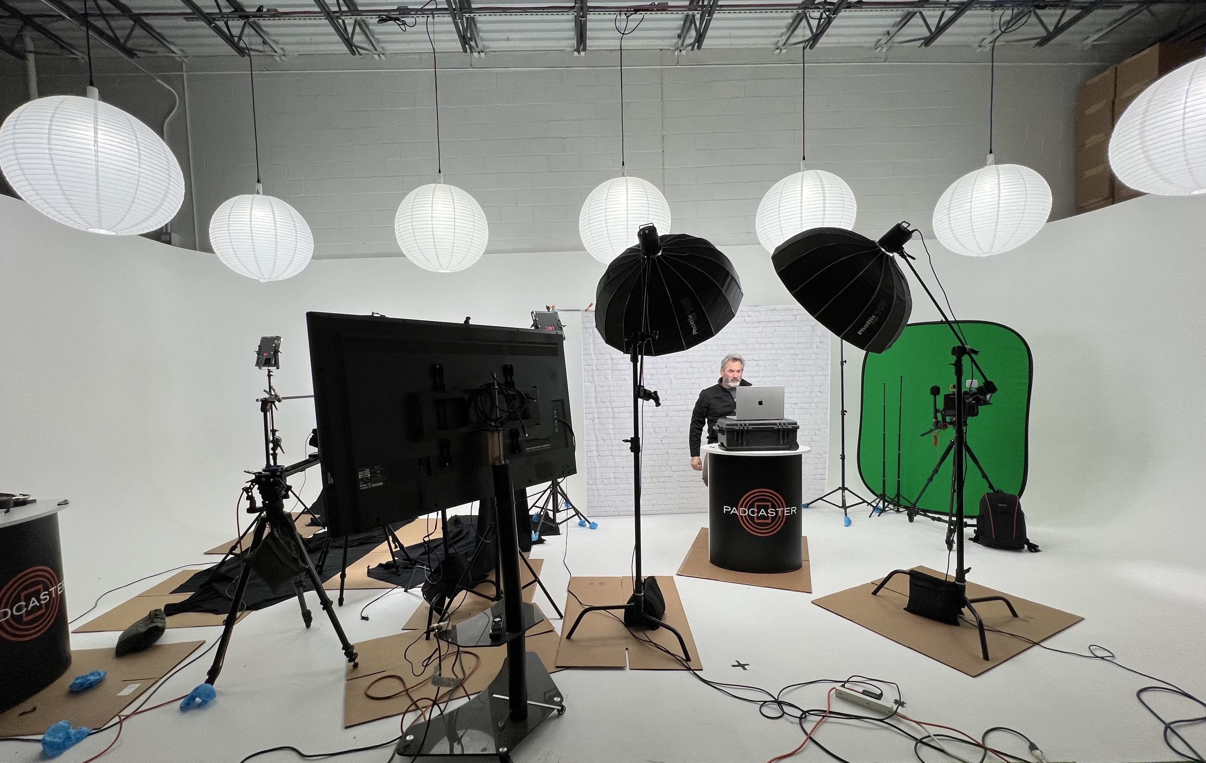 A man stands at a podium with a laptop in a studio setup featuring lights, cameras, and a green screen, surrounded by paper lanterns and various equipment.