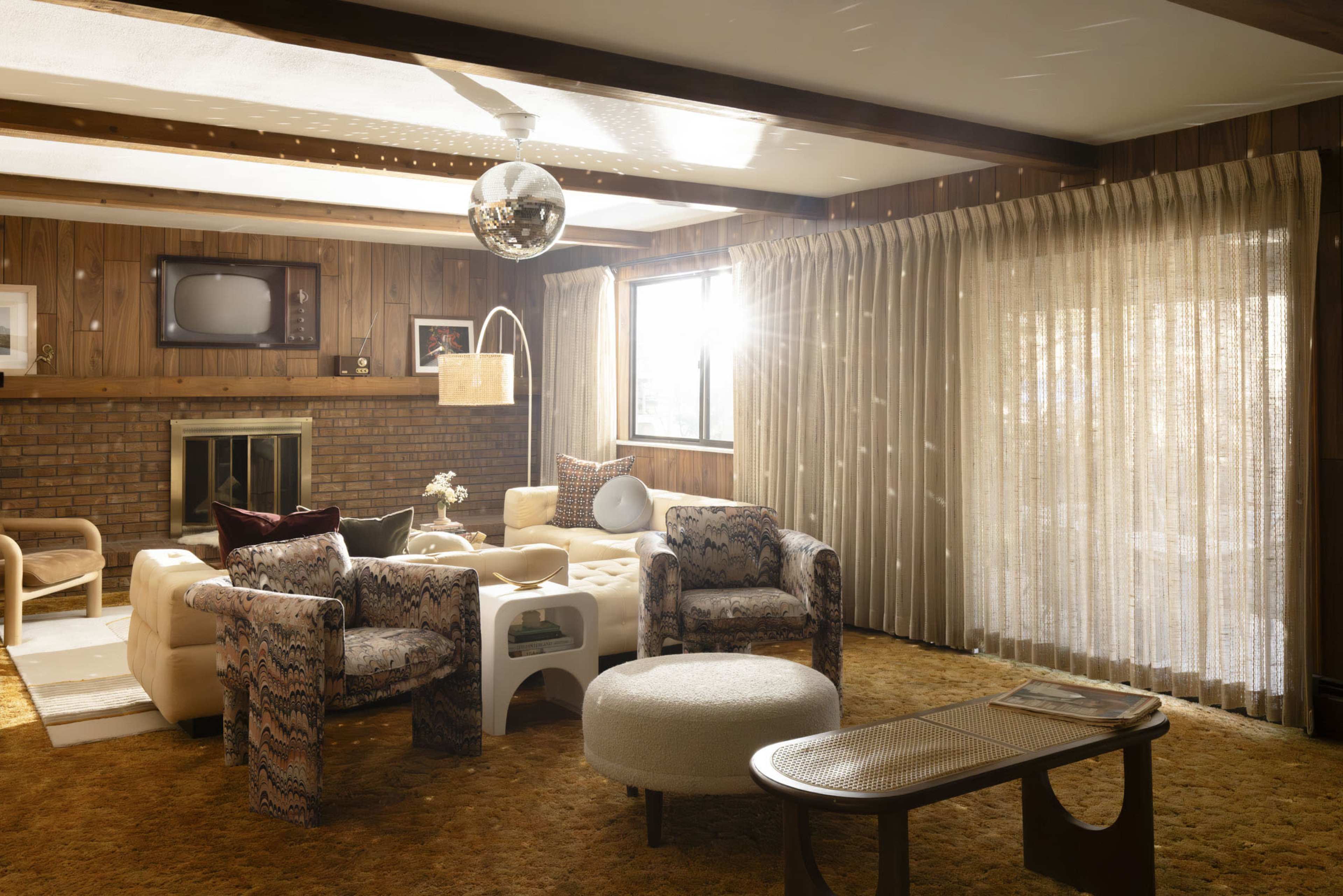 The image shows a retro-style living room with wooden paneling, a brick fireplace, and a large window with sheer curtains letting in sunlight.