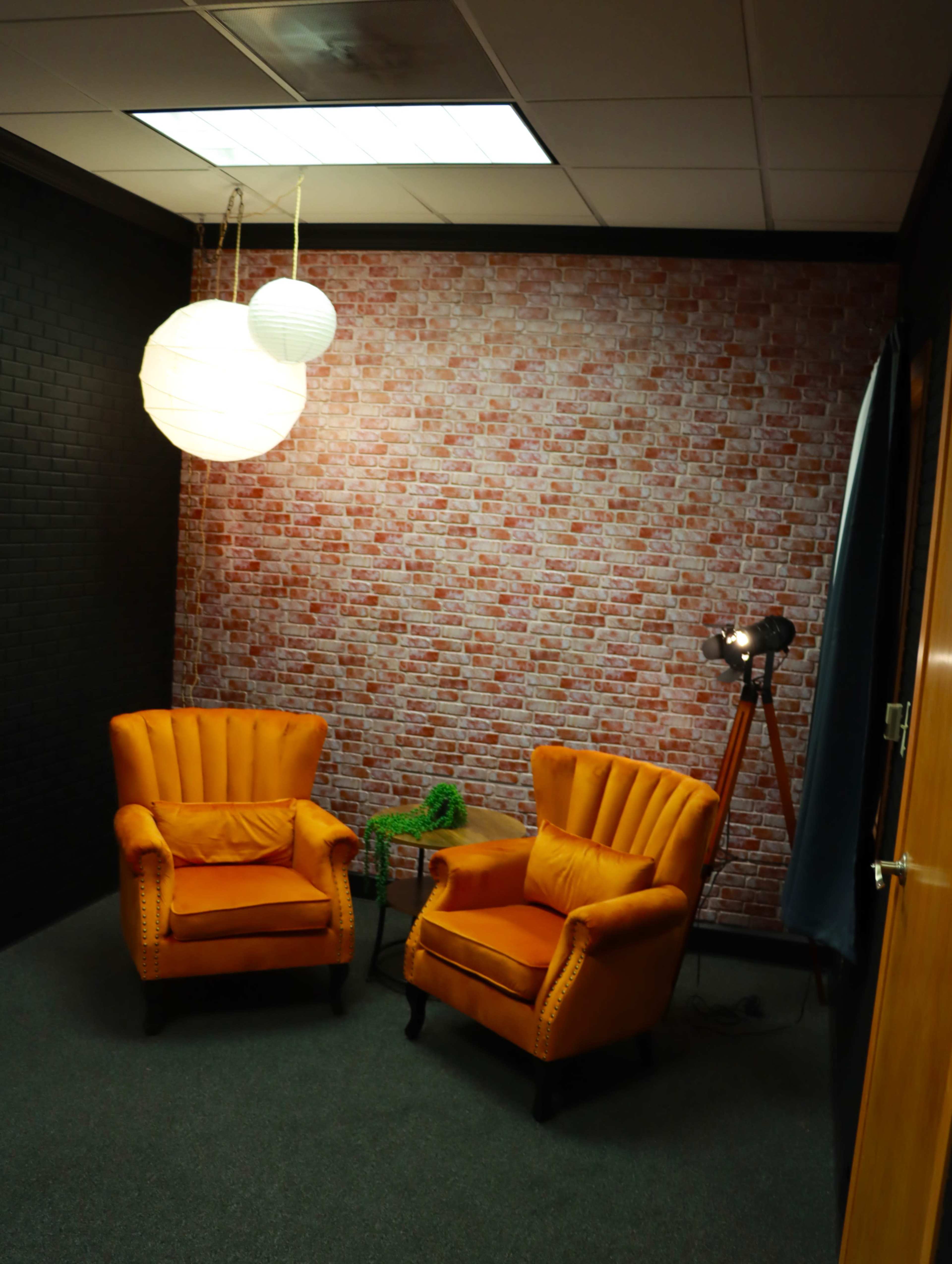 The image shows a small room with two orange armchairs, a wooden side table, a green plant, and hanging light fixtures, all against a brick-patterned wall.