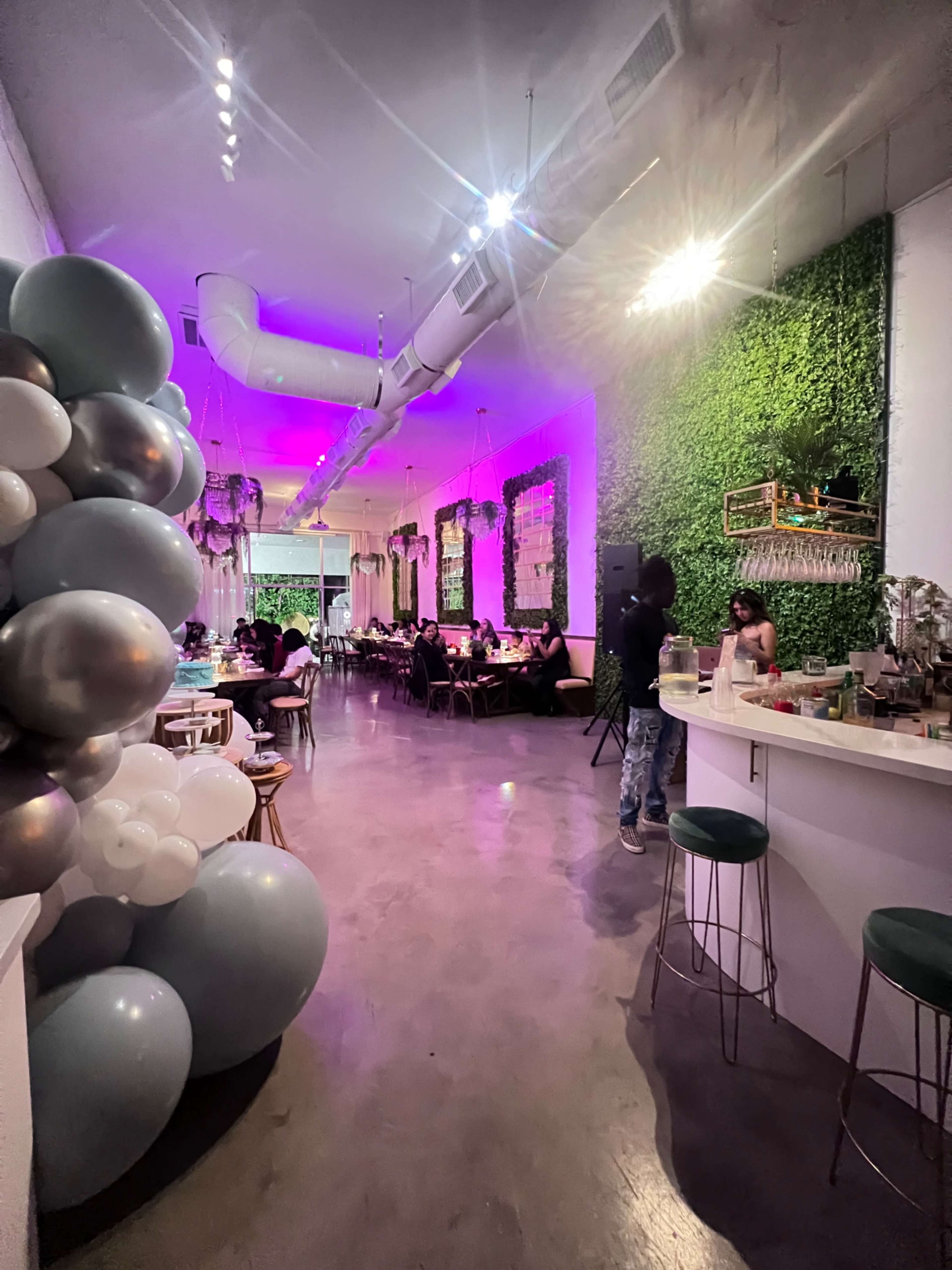 An elegantly decorated restaurant interior with a green wall, a bar area on the right, and tables occupied by diners under purple lighting.