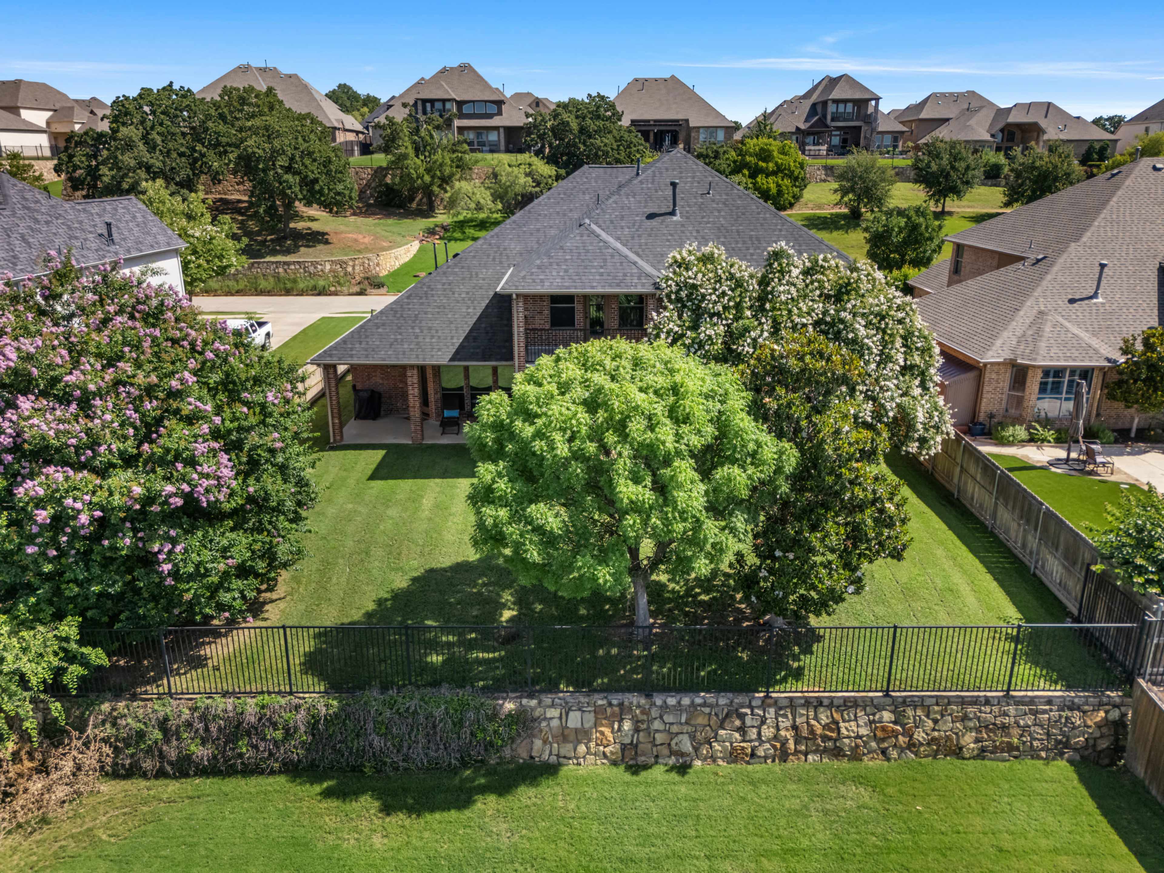 The image shows a residential backyard with a grassy area bordered by a stone wall, featuring several trees and houses in the background.