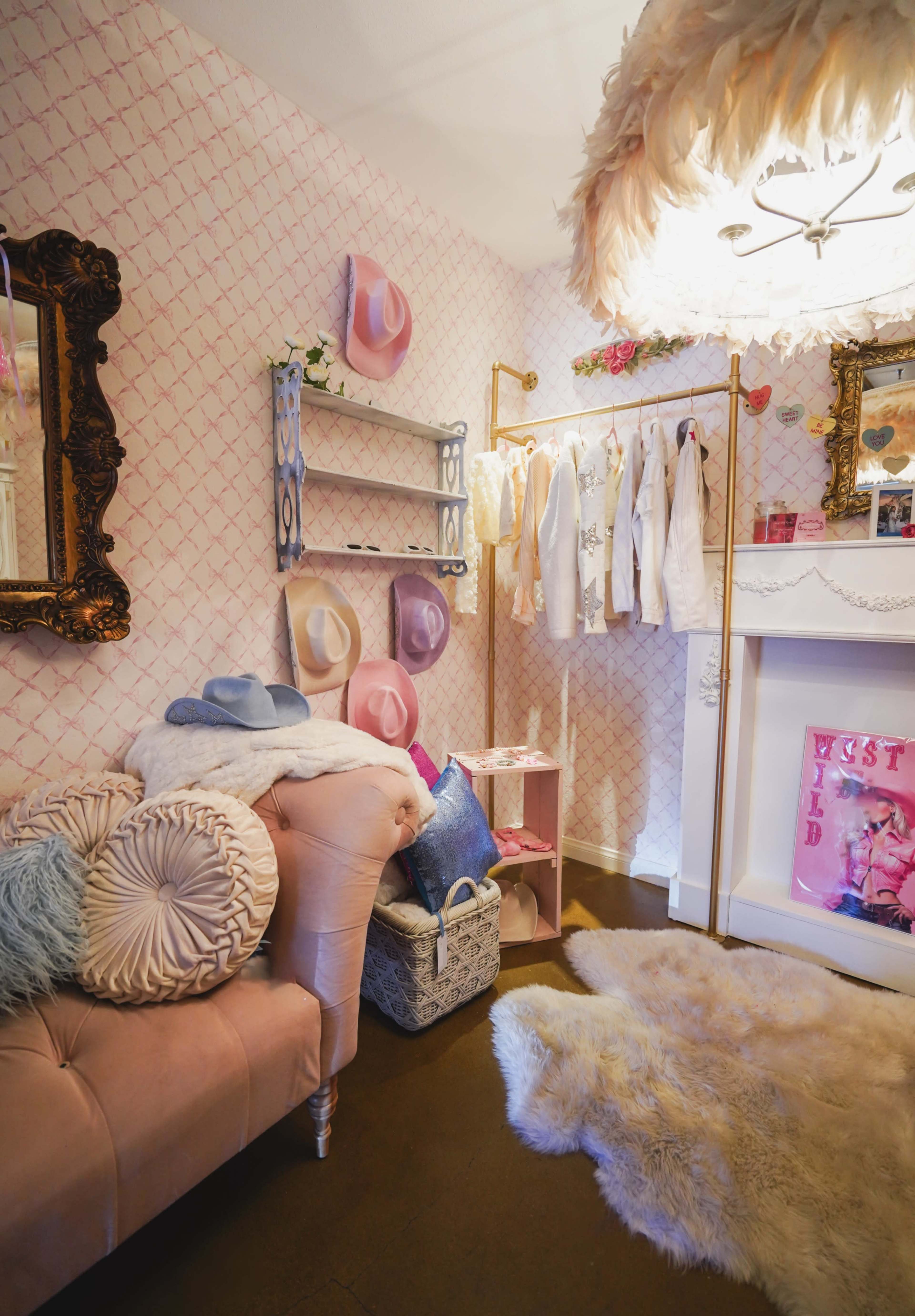 A stylish dressing room with a pink and white patterned wallpaper, a velvet sofa, a clothing rack displaying white garments, and several hats hung on the wall.