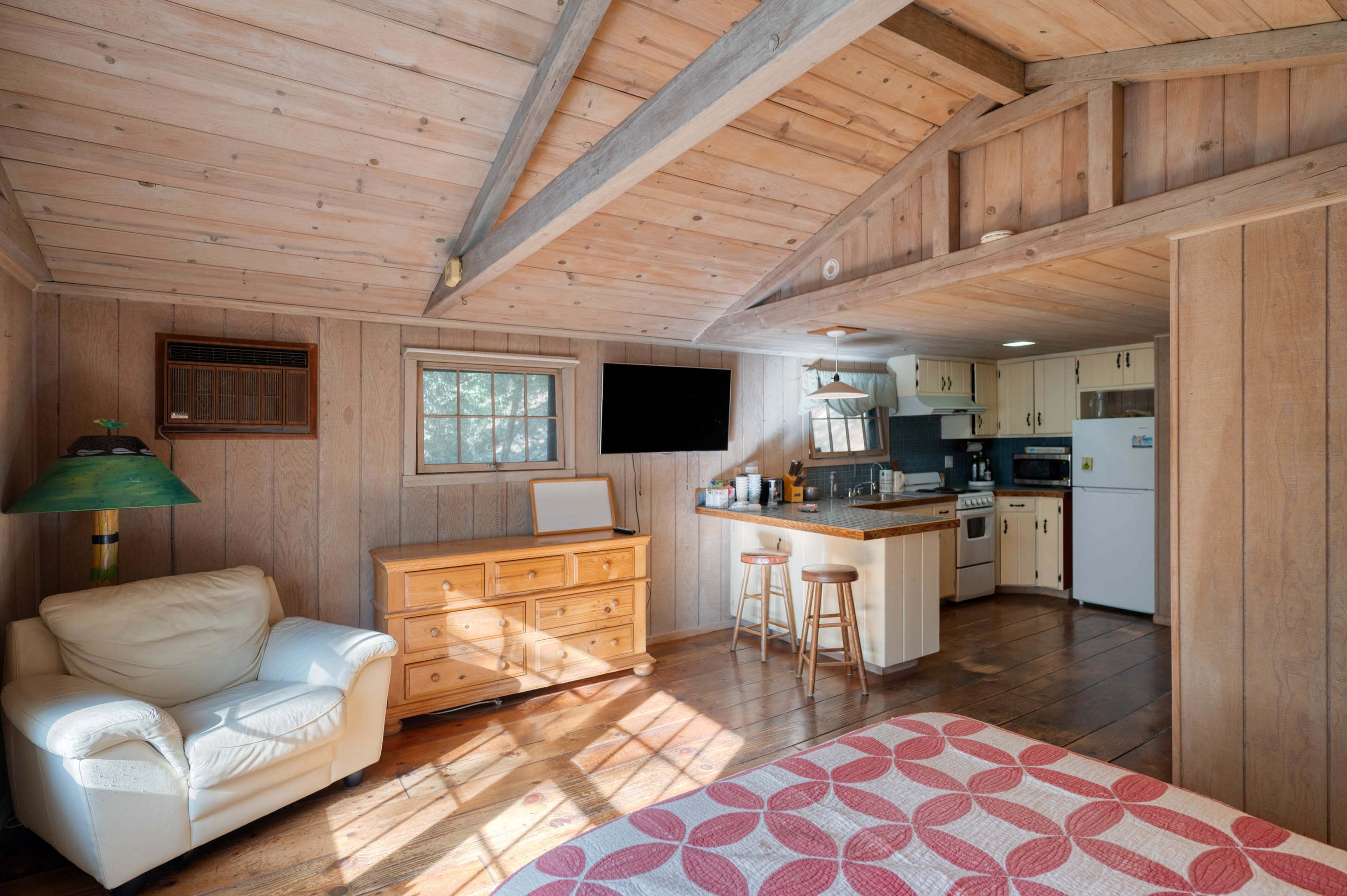 The image depicts a rustic wooden cabin interior featuring a bed, a living area with a white chair, a wooden dresser, and a kitchenette.