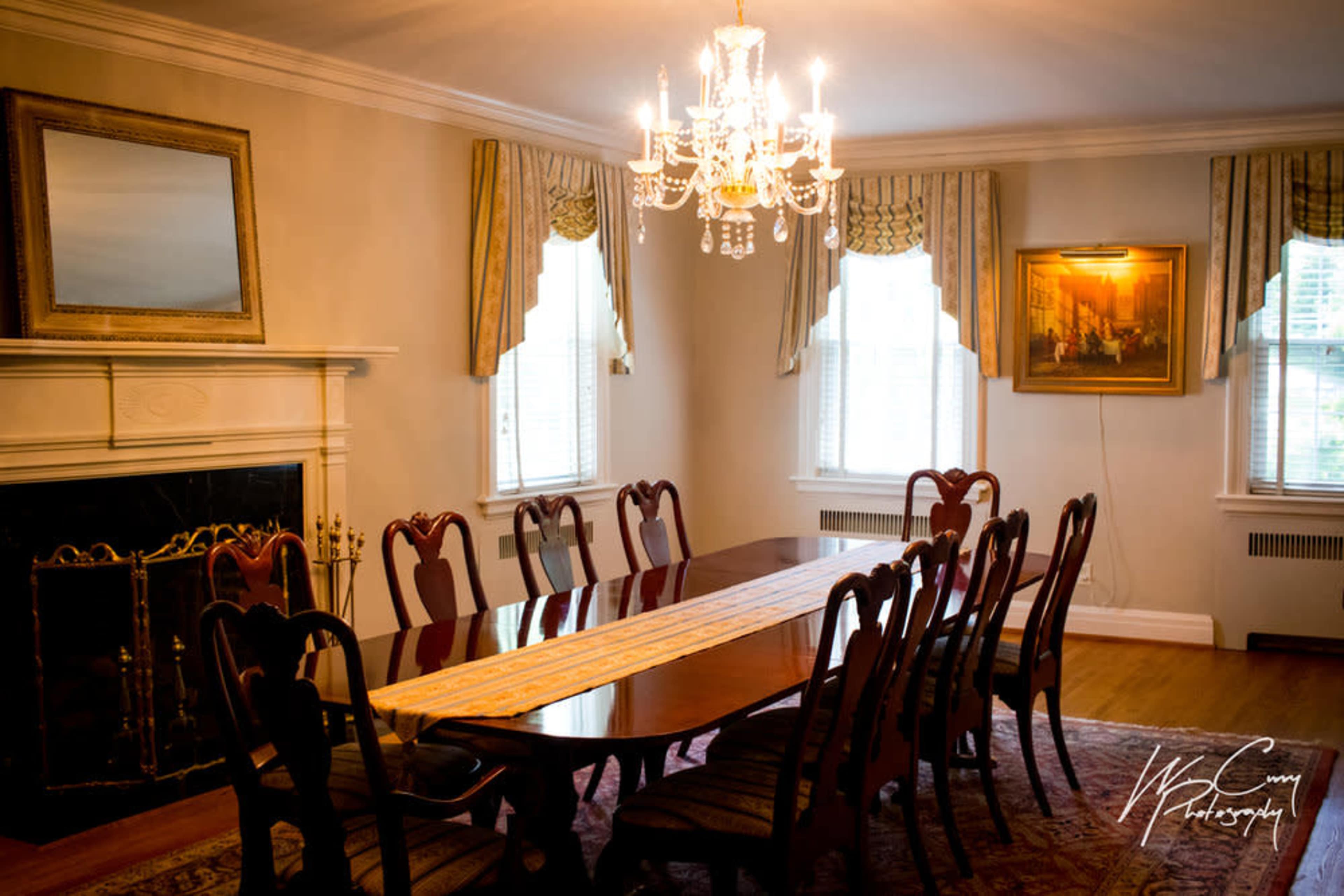 A formal dining room features a long wooden table surrounded by twelve chairs, illuminated by a chandelier and adorned with artwork on the walls.