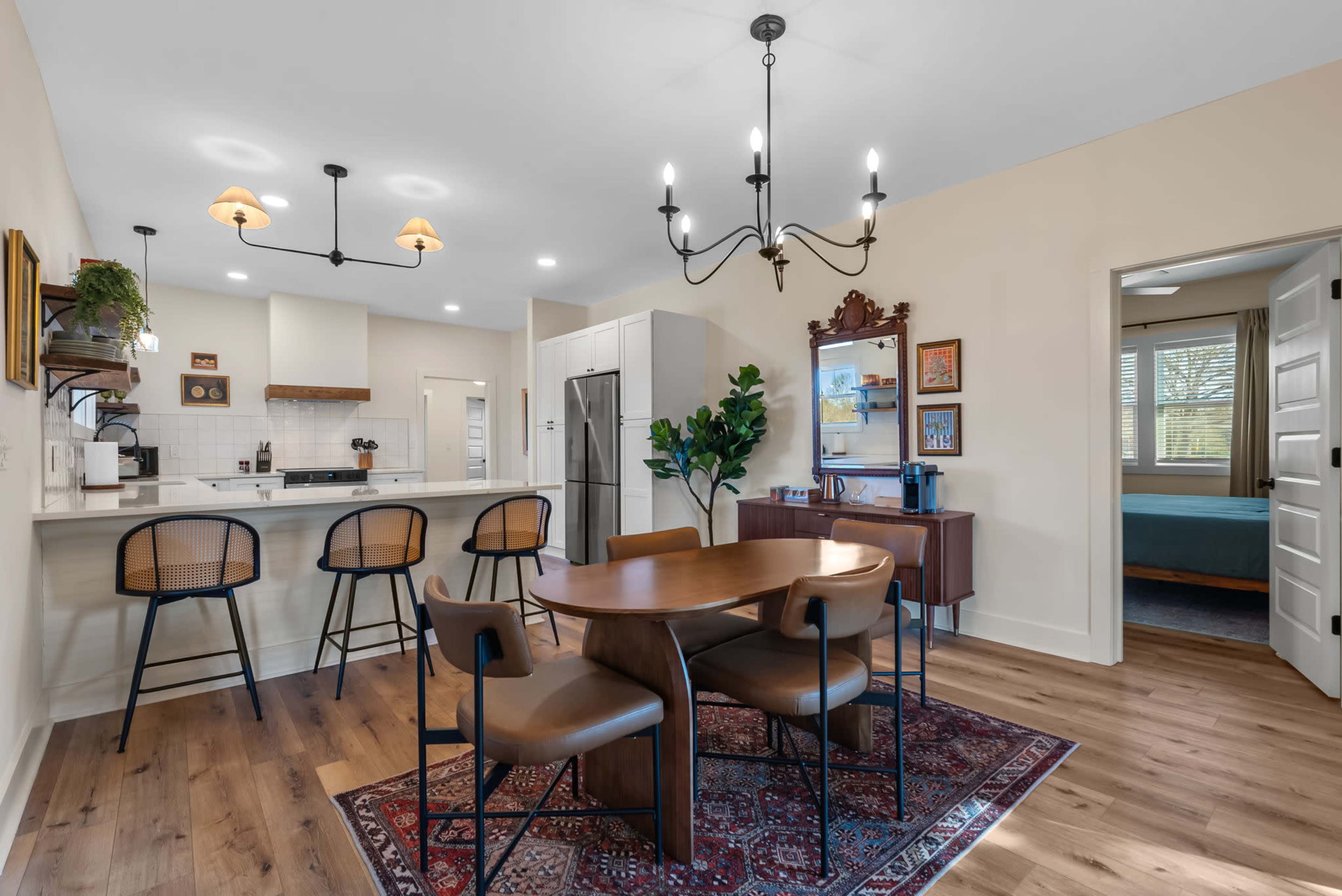 A modern dining area features a wooden table with four chairs, complemented by a chandelier and an open kitchen in the background.