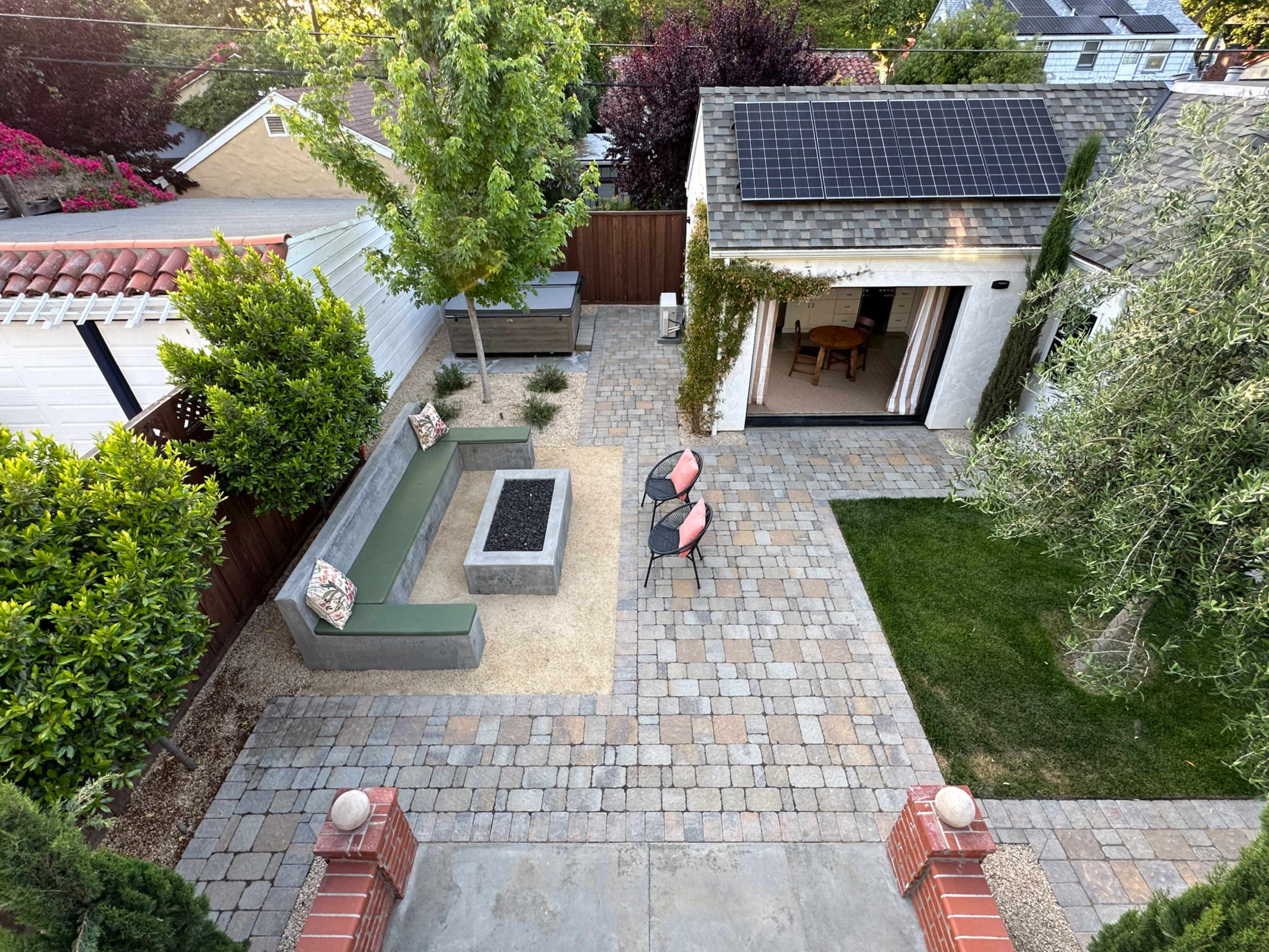 The image shows a landscaped backyard with a stone-paved patio, a fire pit seating area, and a small shed featuring solar panels.
